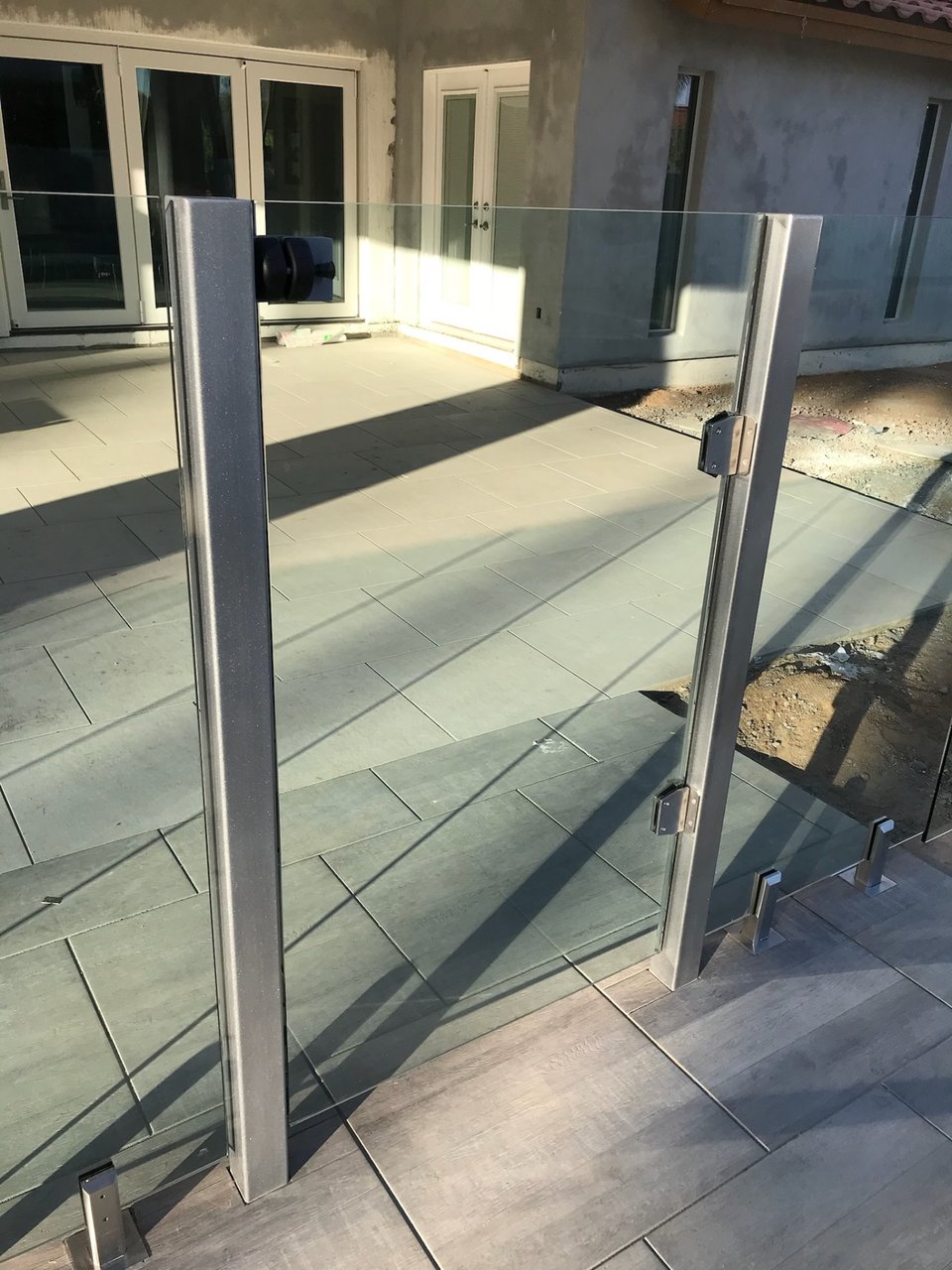 Metal gate posts and a hinged frameless glass gate panel beside paver decking — close detail of the spigot, hinge, and latch hardware.