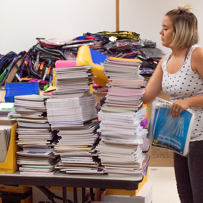 Volunteer organizing tall stacks of notebooks beside a pile of backpacks at the staging area