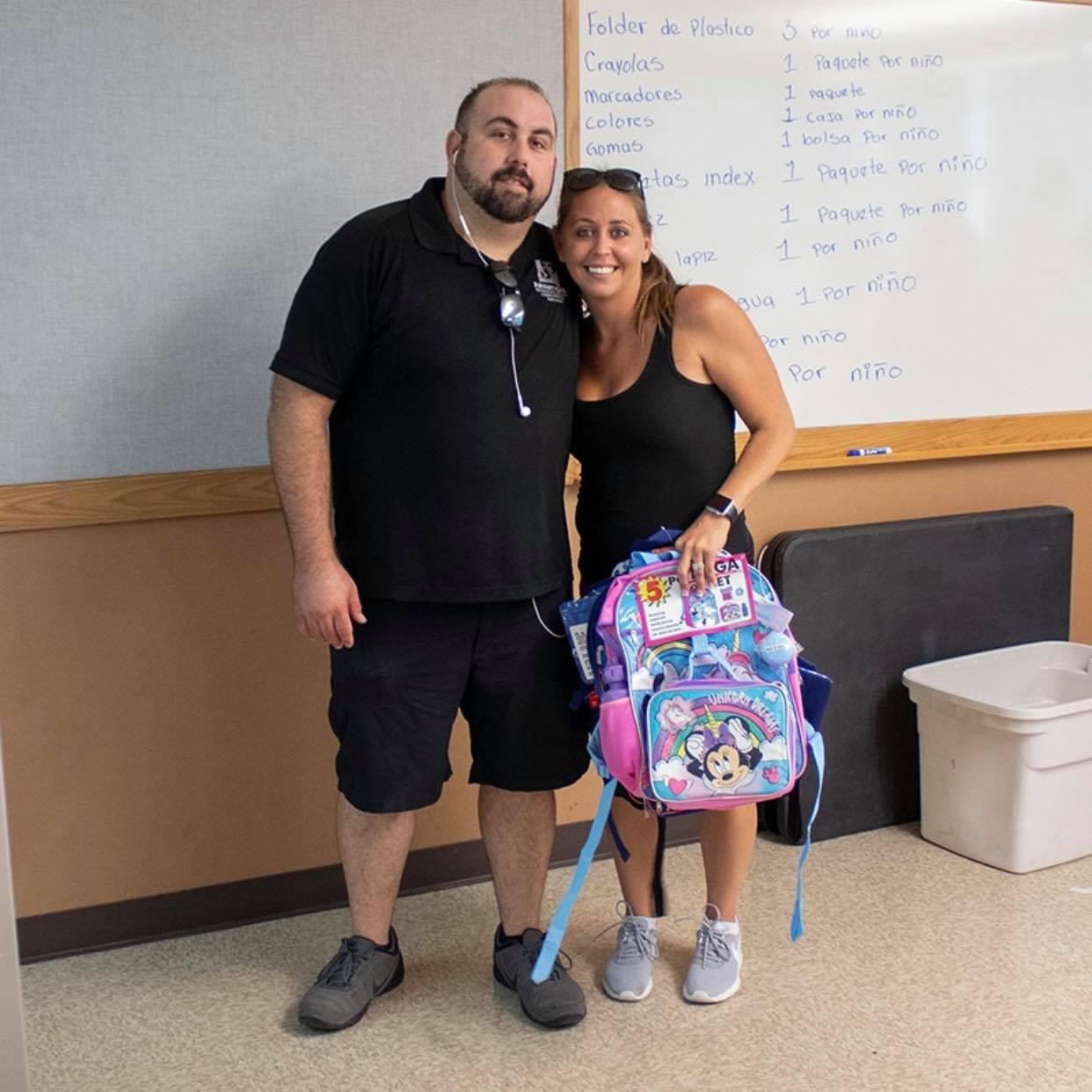 Anthony from AE Outdoor Living with a volunteer holding a Minnie Mouse backpack in front of a Spanish supply list on a whiteboard