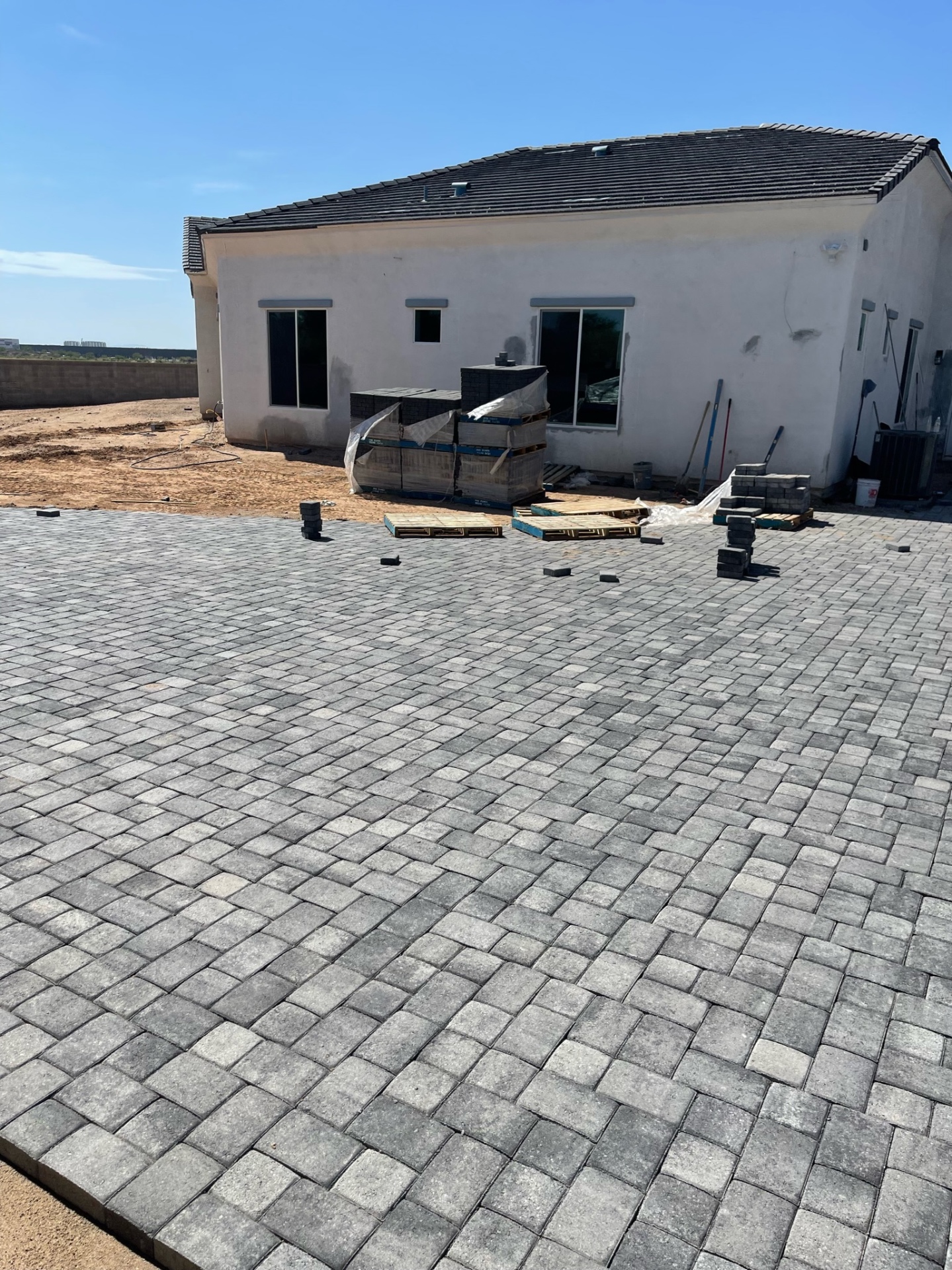 Waddell (Surprise-area) rear charcoal paver patio spanning the back of the stucco home with staged paver bundles ready for final cuts and edge completion by AE Outdoor Living