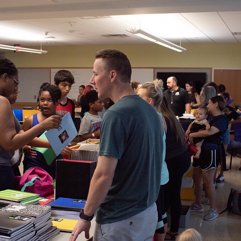 Volunteer in an AE Outdoor Living polo handing a folder of school supplies to a family