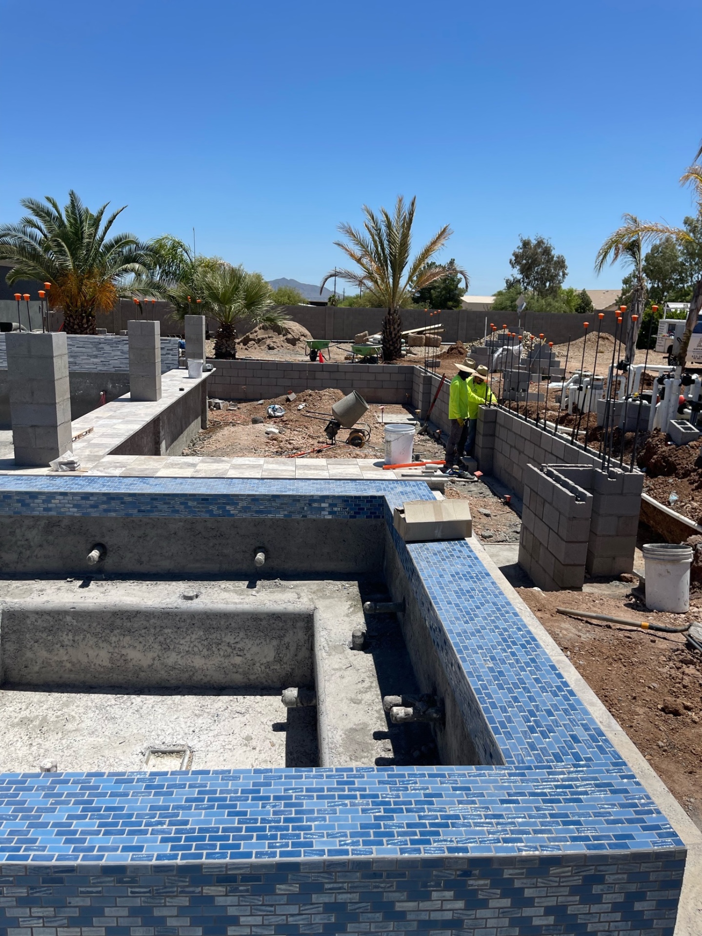 Wide jobsite view with raised tiled spa, pool shell, perimeter walls, and crew member finishing block beside the equipment pad