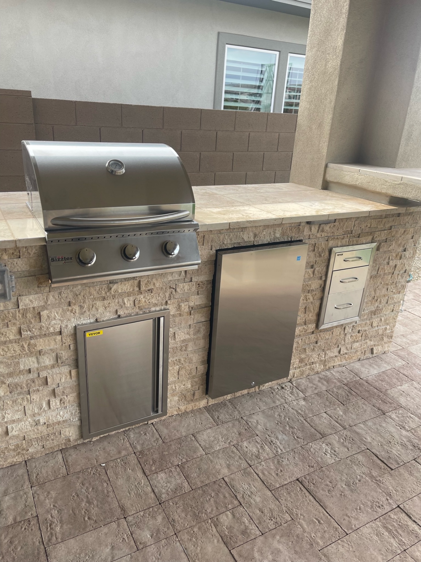 Peoria covered outdoor kitchen with stacked-stone BBQ island, travertine tile counter, Sizzler stainless grill, drawer stack and beverage fridge by AE Outdoor Living