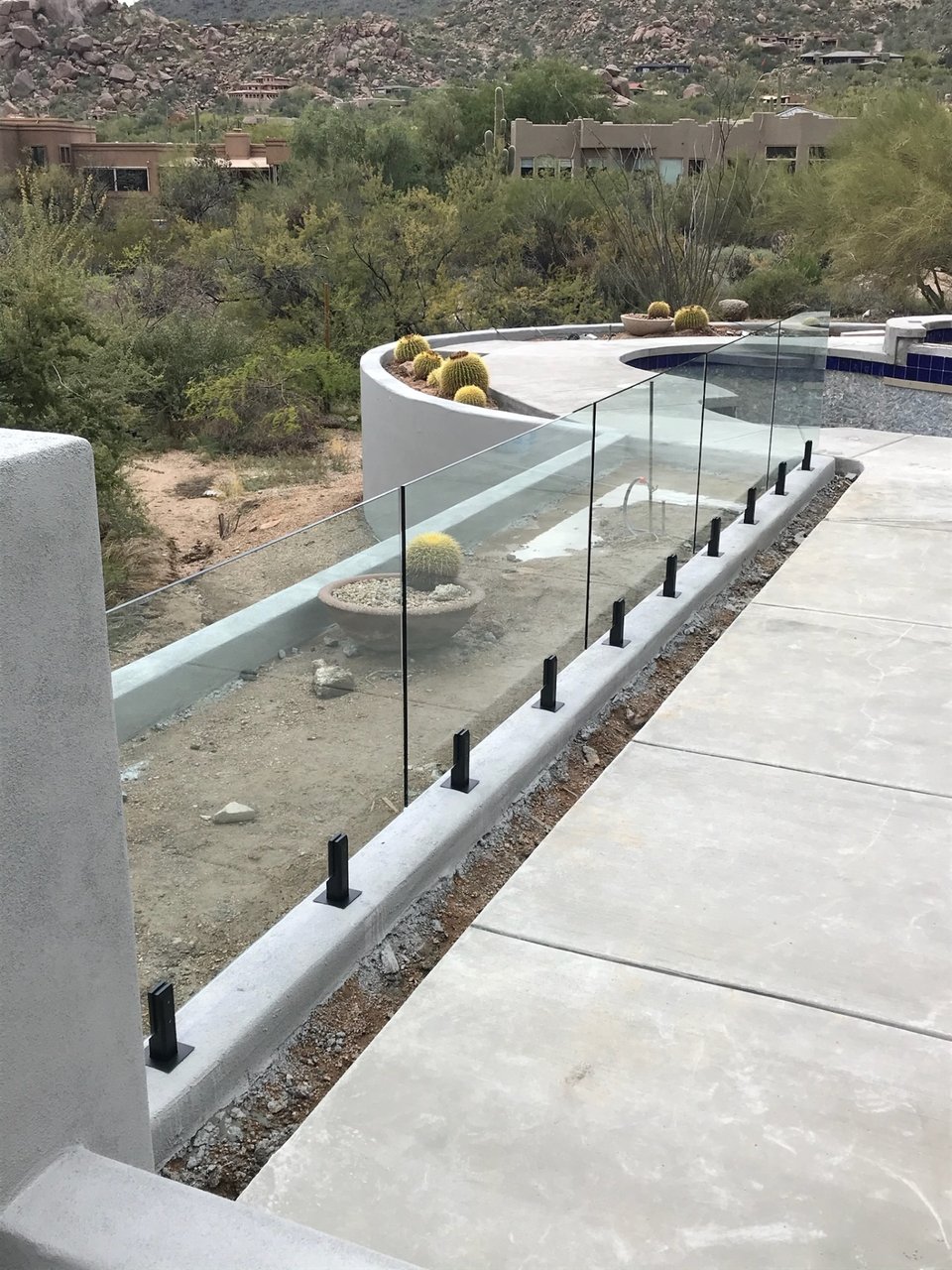 Curved run of glass fence panels on a poured concrete curb, with a boulder hillside and barrel cactus planters beyond.