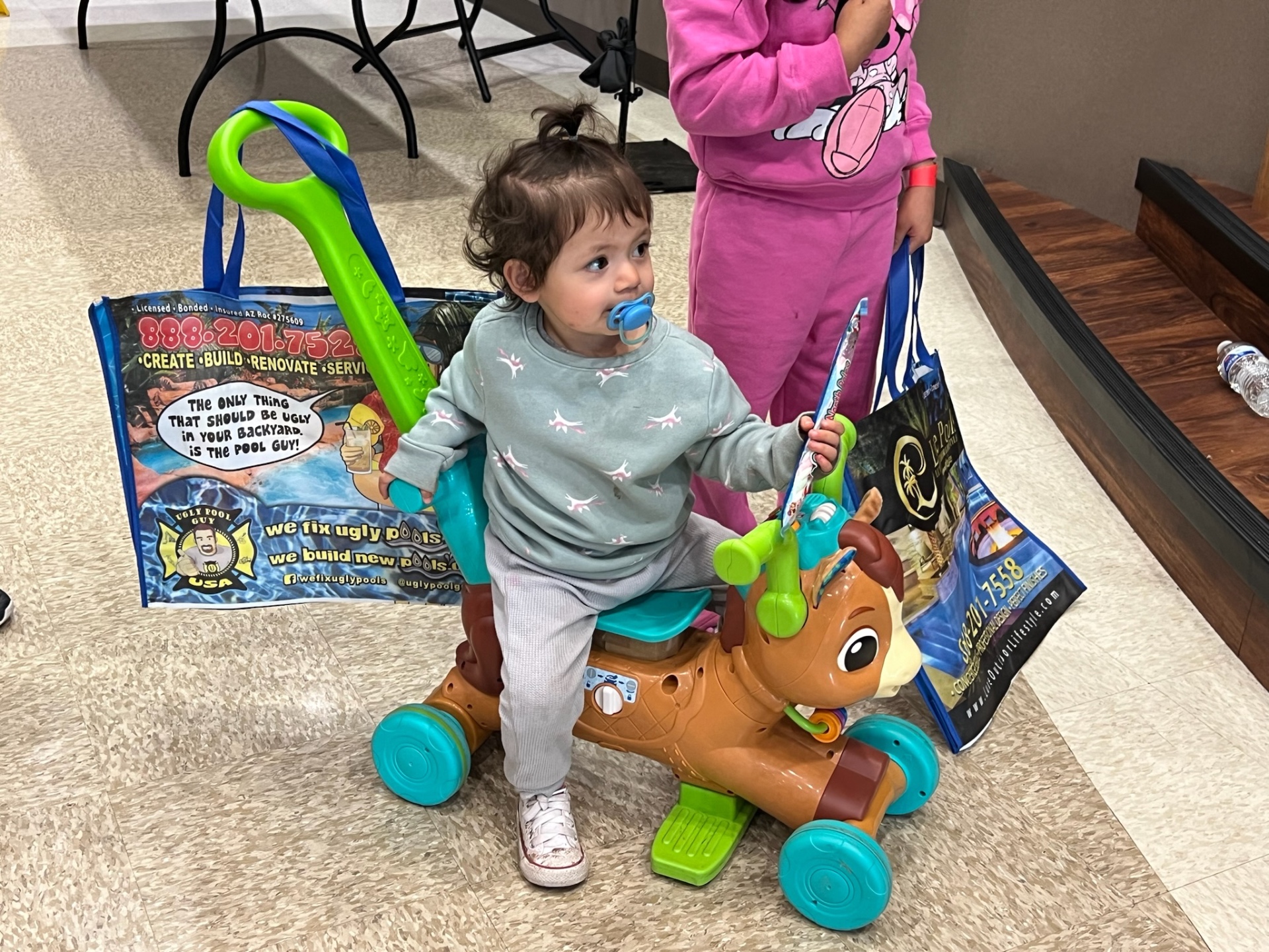Toddler riding a toy horse next to AE Outdoor Living Ugly Pool Guy and Luxe Pools gift bags at Shop With a Cop