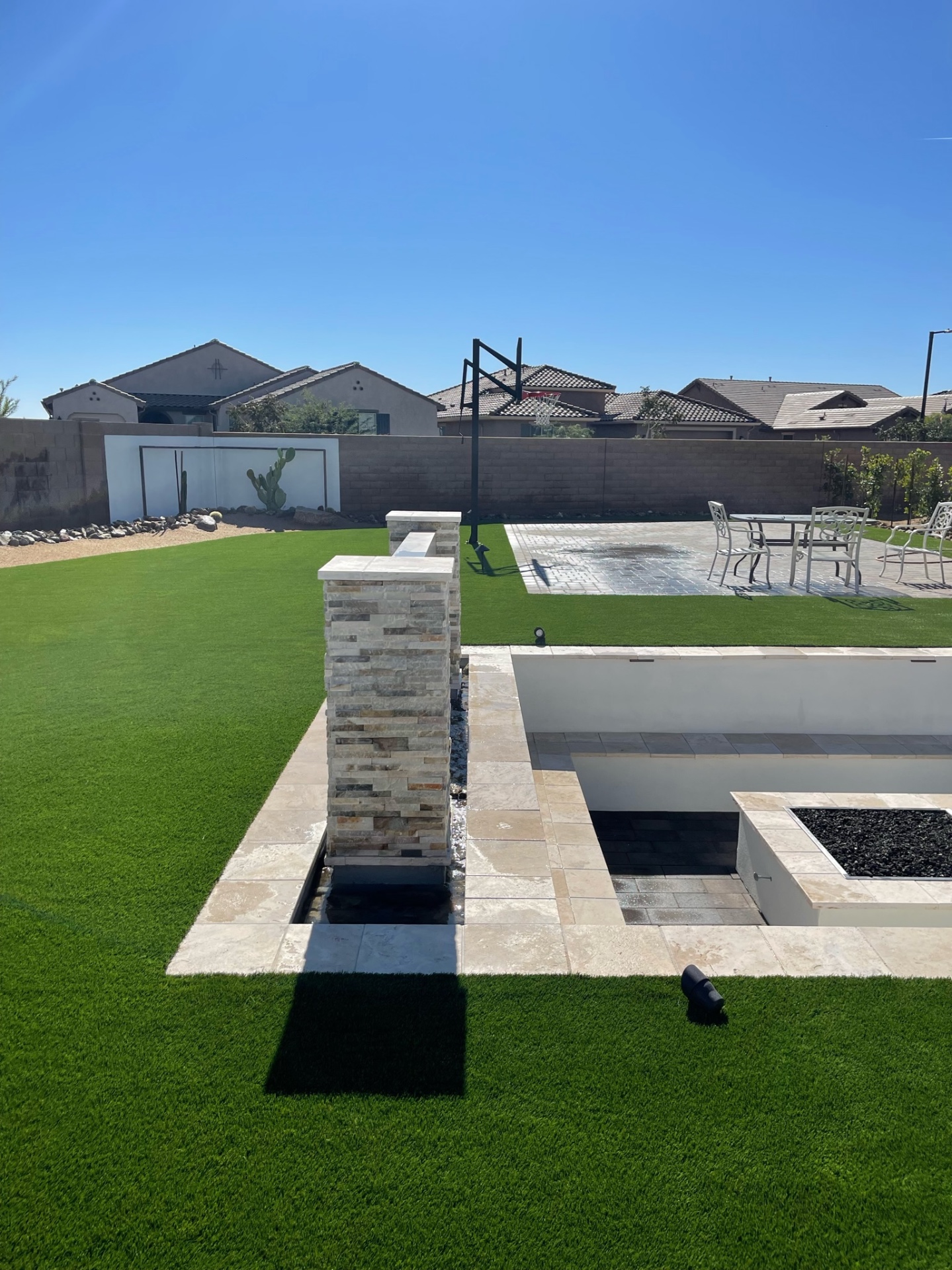 Goodyear backyard sunken firepit with travertine seat walls, stacked-stone water feature column and paver sport court beyond a fresh turf lawn