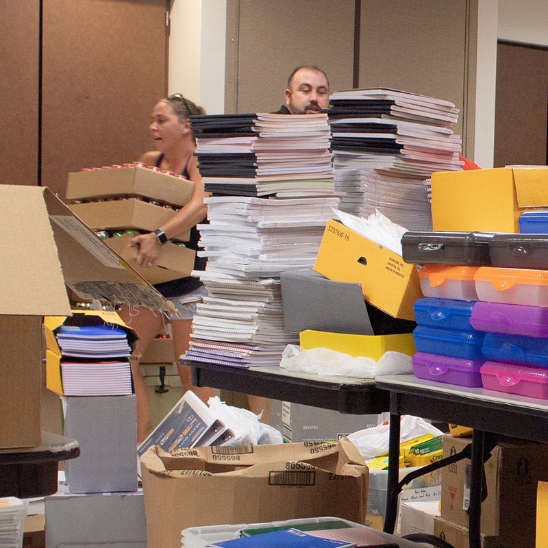 Volunteers moving boxes of supplies past towering stacks of notebooks at the community center