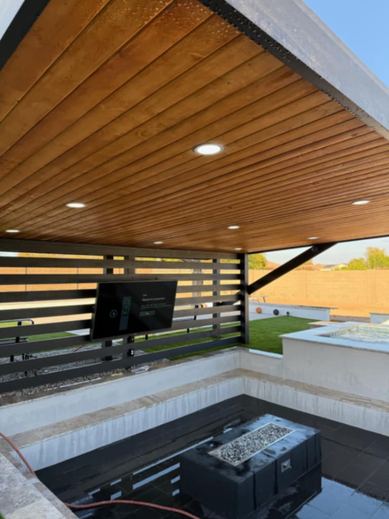 Wide interior view of the black pergola — wood slat wall, cedar ceiling, TV, and sunken fire lounge below.