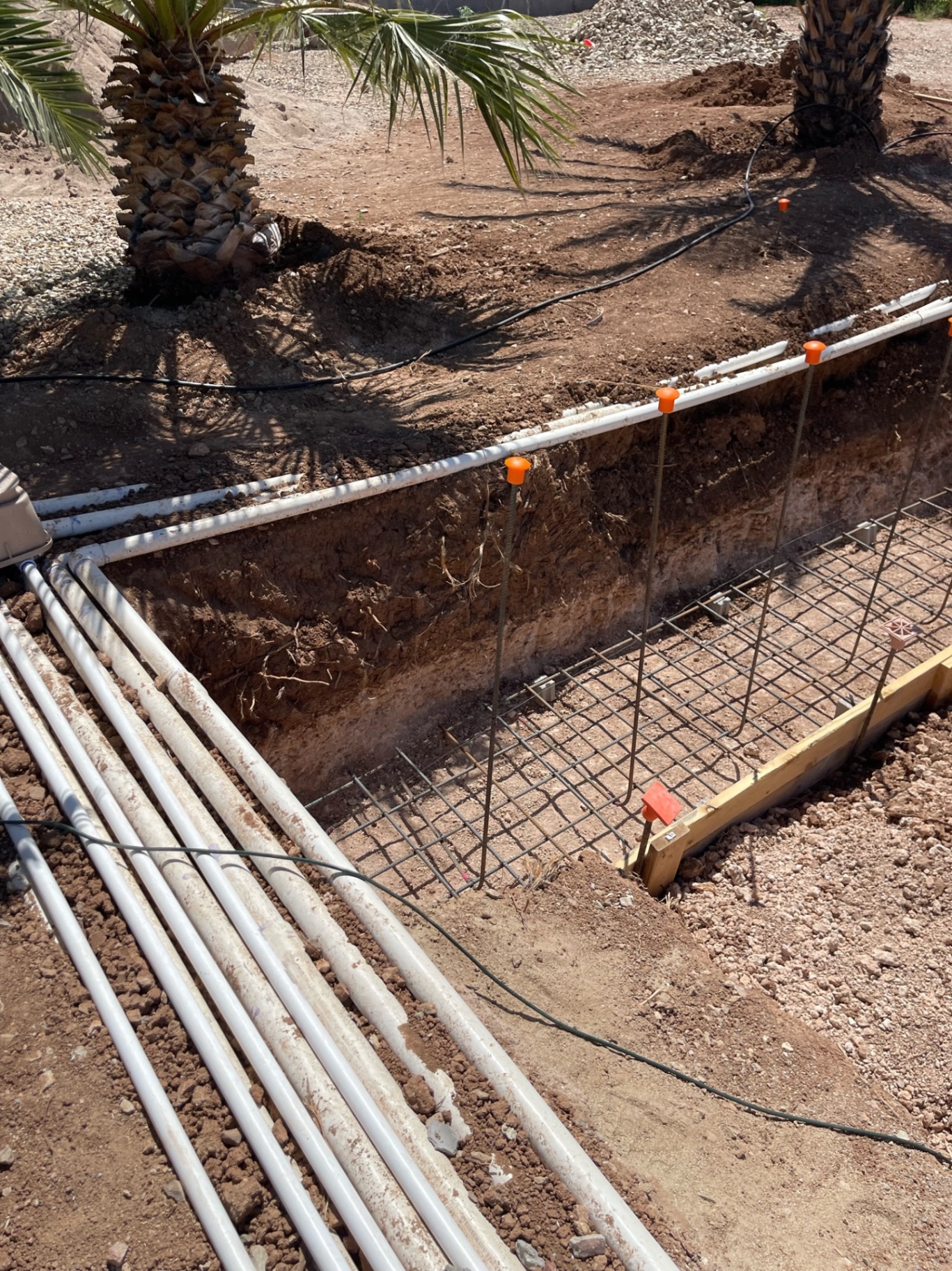 Open footing trench with vertical rebar, dirt stockpiles, and multiple PVC runs stacked beside the excavation