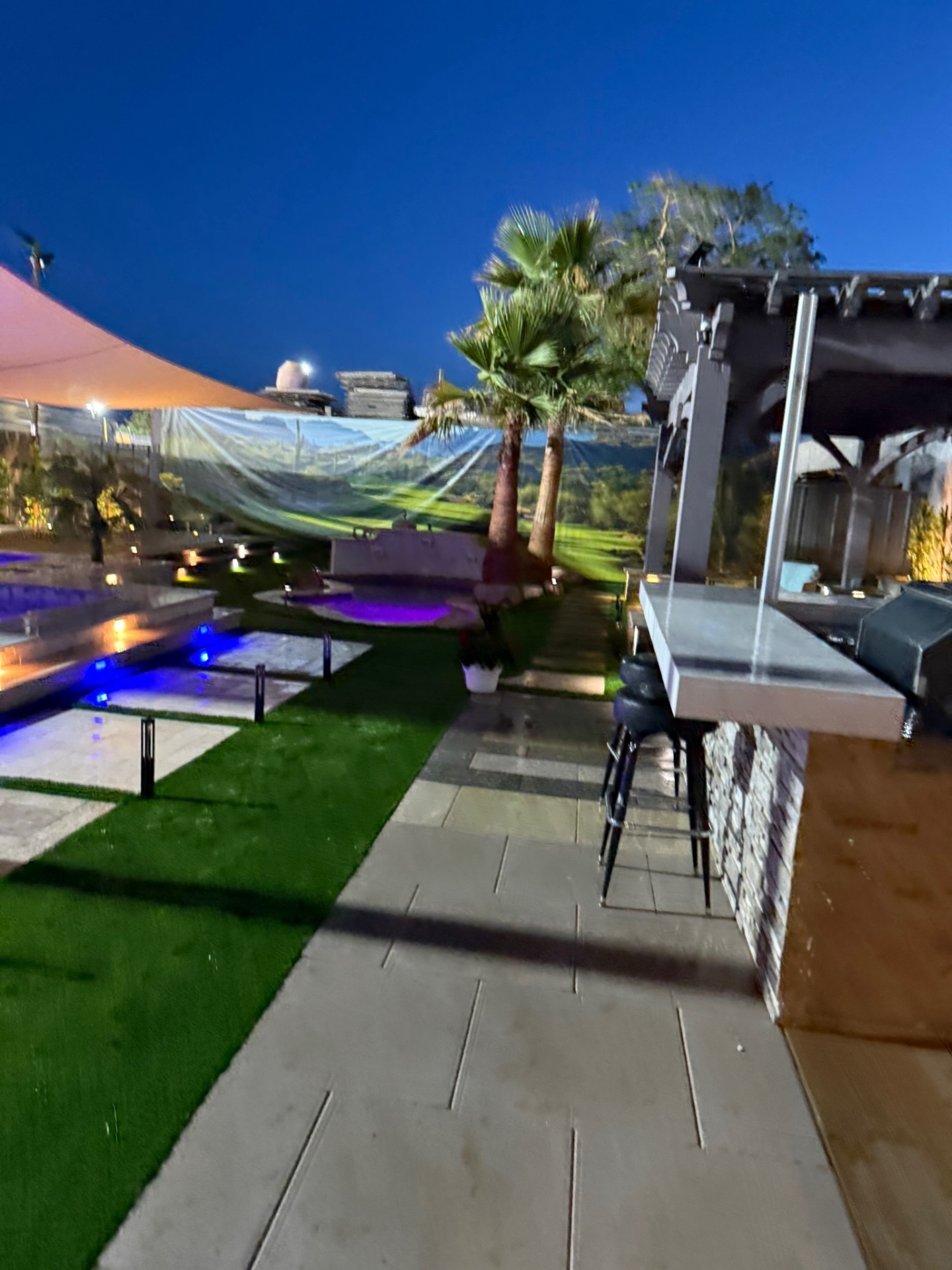 Wide view of display yard with bar seating, pool, and shade sails at dusk