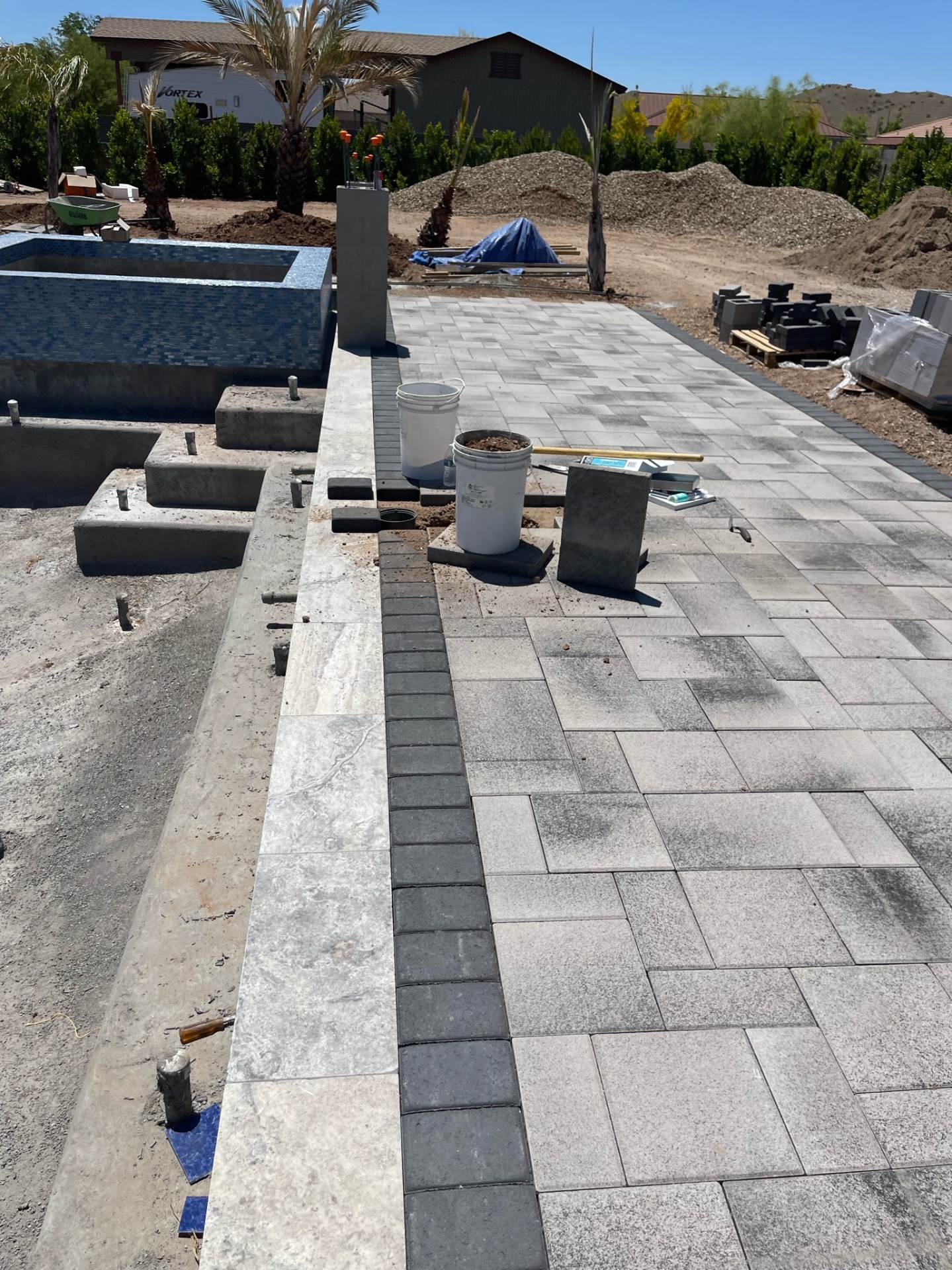 Nearly finished large-format paver deck with dark border band, raised spa and stacked-stone wall visible at the far side