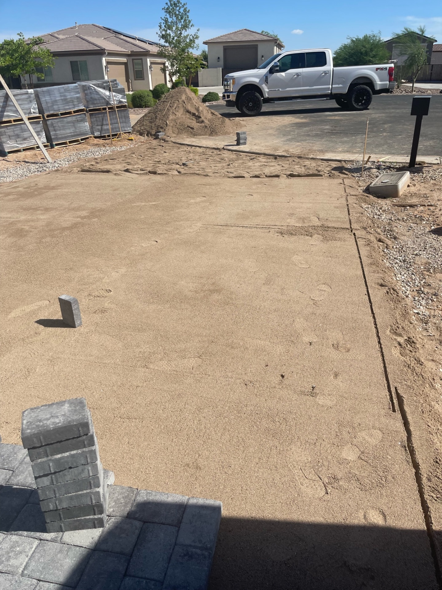 Waddell (Surprise-area) front-yard paver install in progress — compacted ABC base with 1-inch screeded sand bed and staged charcoal paver pallets on driveway approach by AE Outdoor Living
