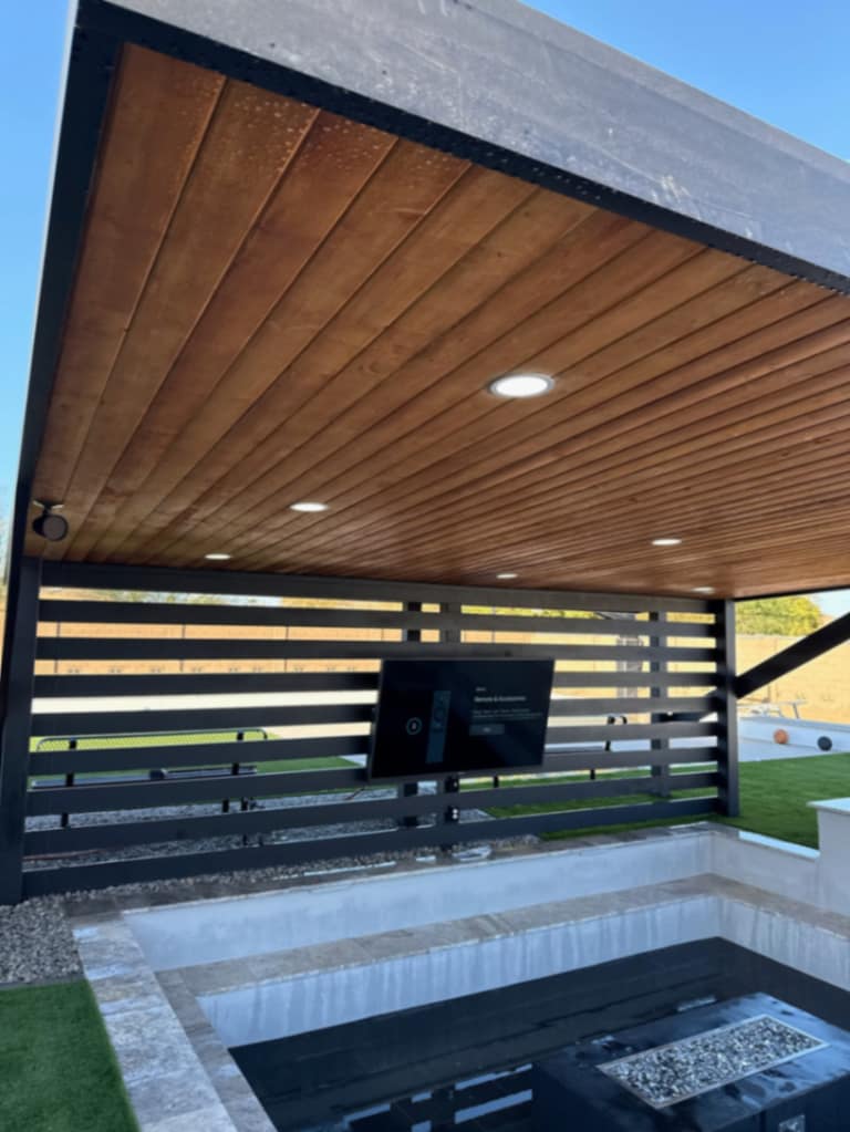 Modern black steel pergola with warm cedar plank ceiling, recessed lighting, mounted TV, and horizontal privacy slats.