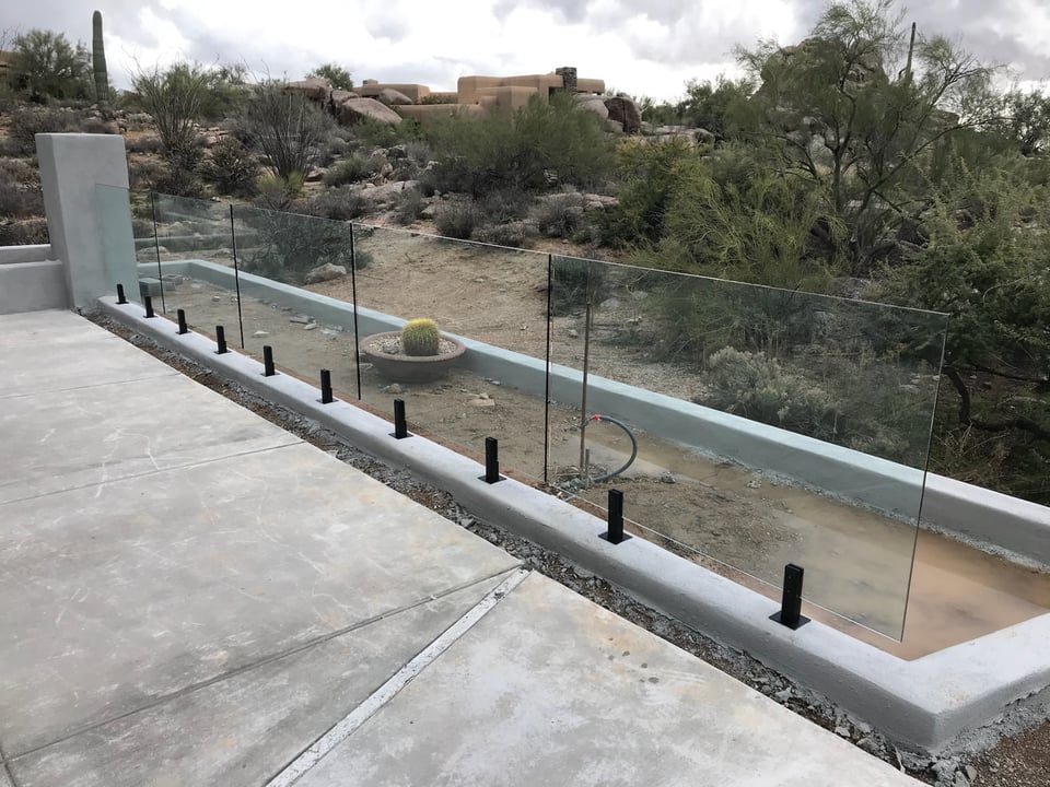 Straight run of glass fence panels along a Sonoran Desert pool deck, with saguaros and Pueblo-style homes in the distance.