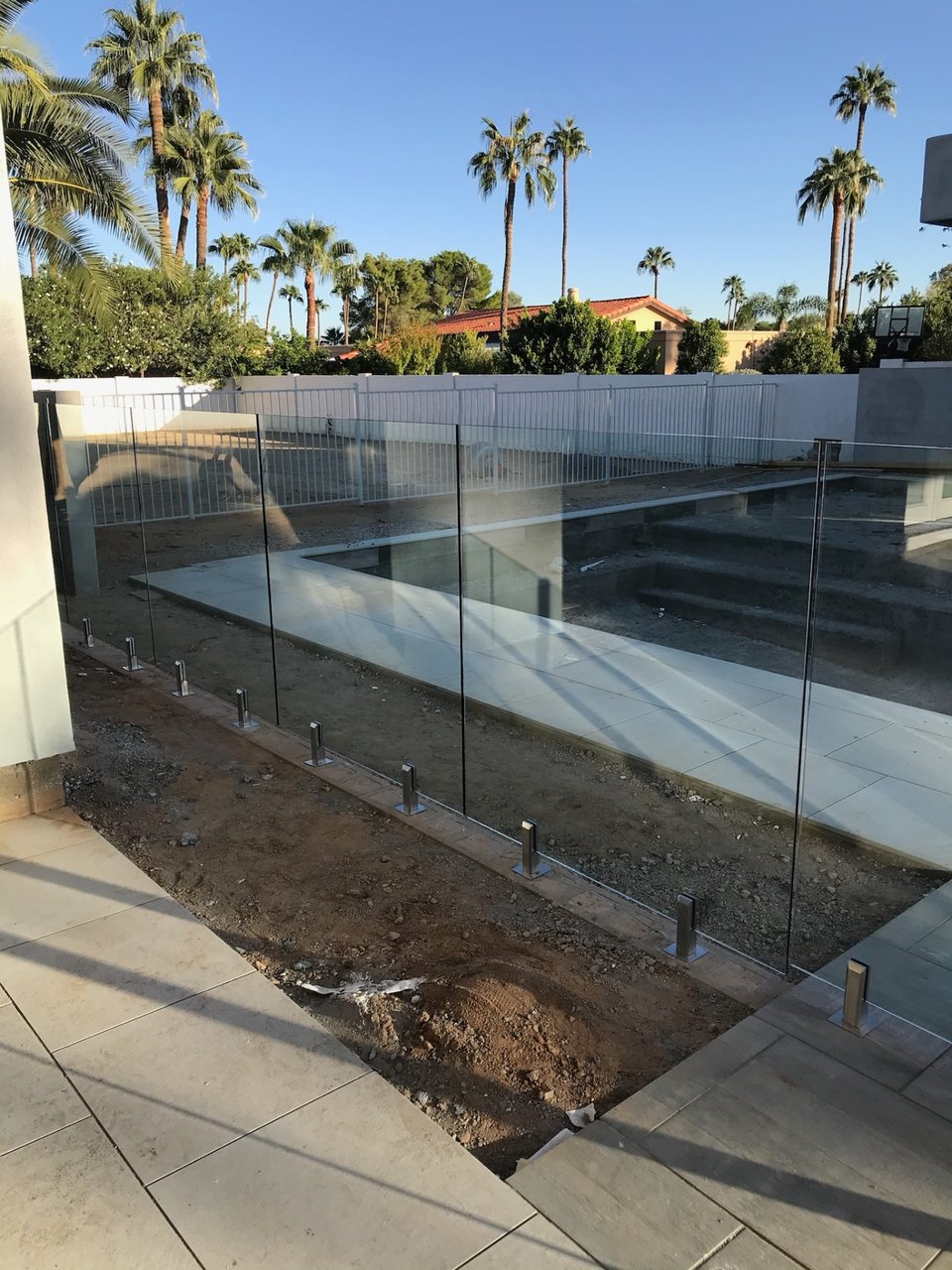 Glass fence panels on spigots along a pool deck surface, with palms and mature landscape behind.