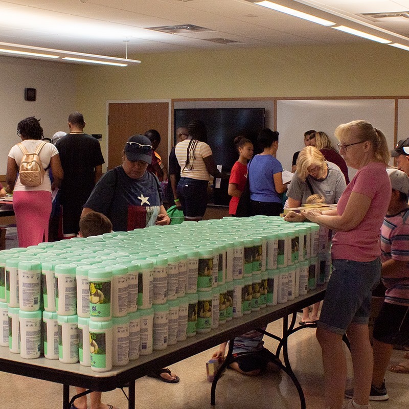 Families lined up around tables stacked with disinfecting wipes at the backpack drive