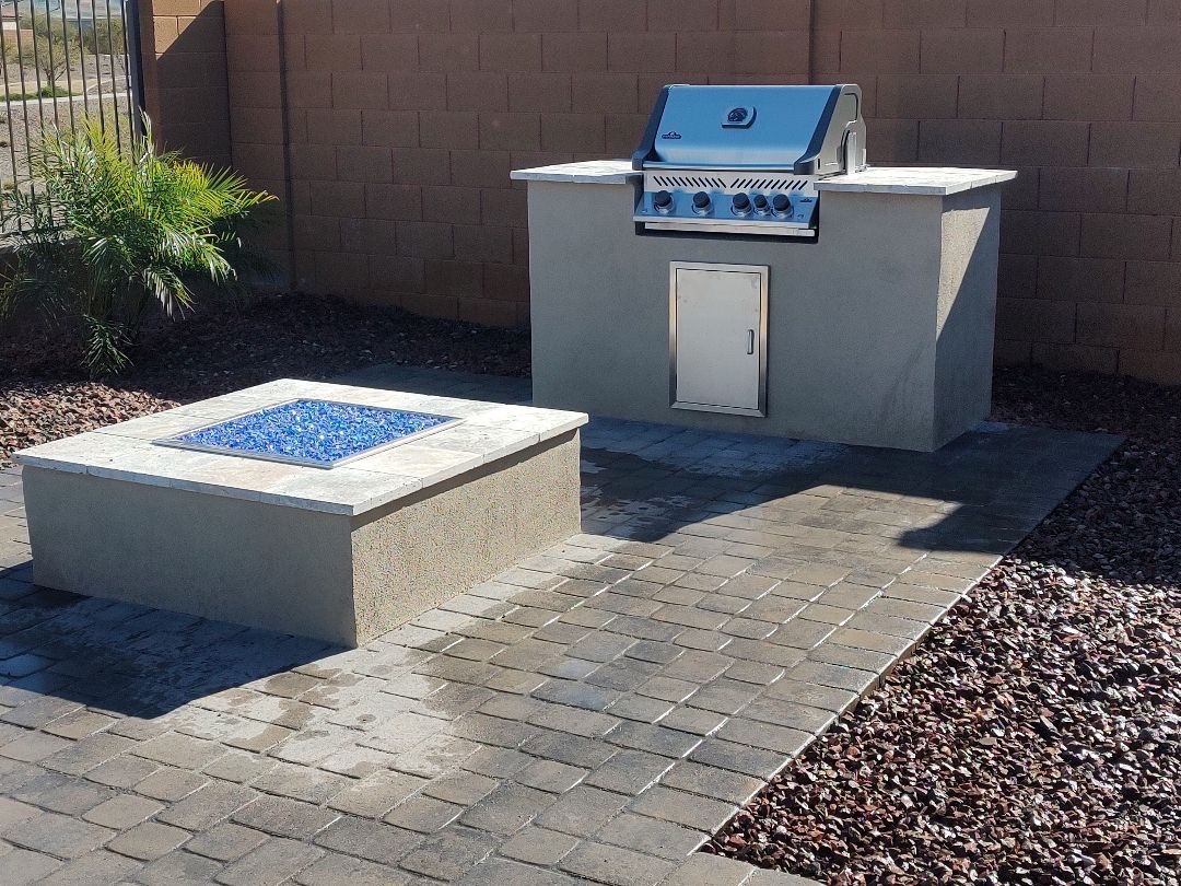 Vistancia (Peoria) backyard paver patio with stucco outdoor kitchen island housing Napoleon built-in grill, travertine counter and matching stucco fire pit with blue fire glass by AE Outdoor Living