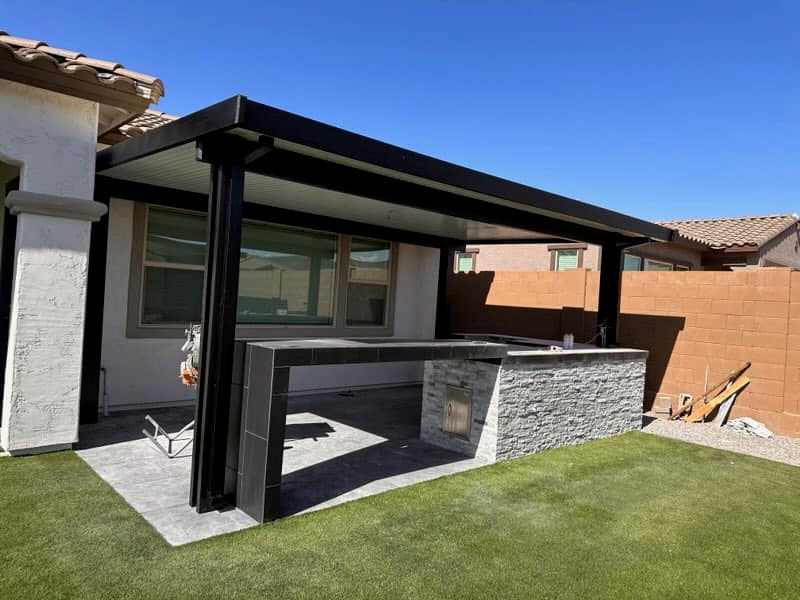 Blackout aluminum patio cover shading a stacked-stone outdoor kitchen and porcelain-tile counters.