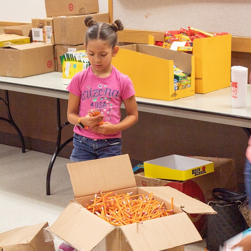 Young girl in an Arizona USA shirt grabbing a handful of pencils from a box at the supply table