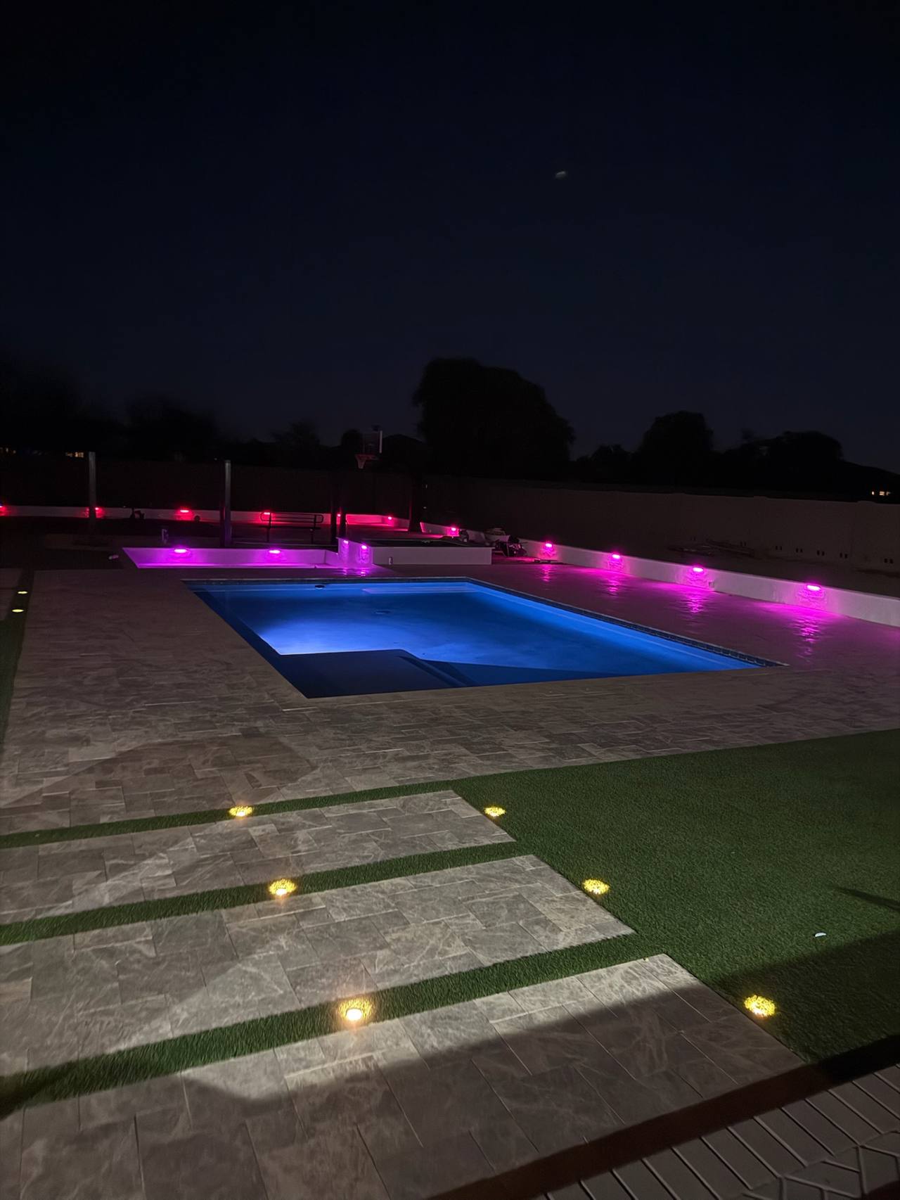 Rectangle pool with blue LED, pink accent wall lights, travertine deck, and turf-and-paver ribbon walkway at night