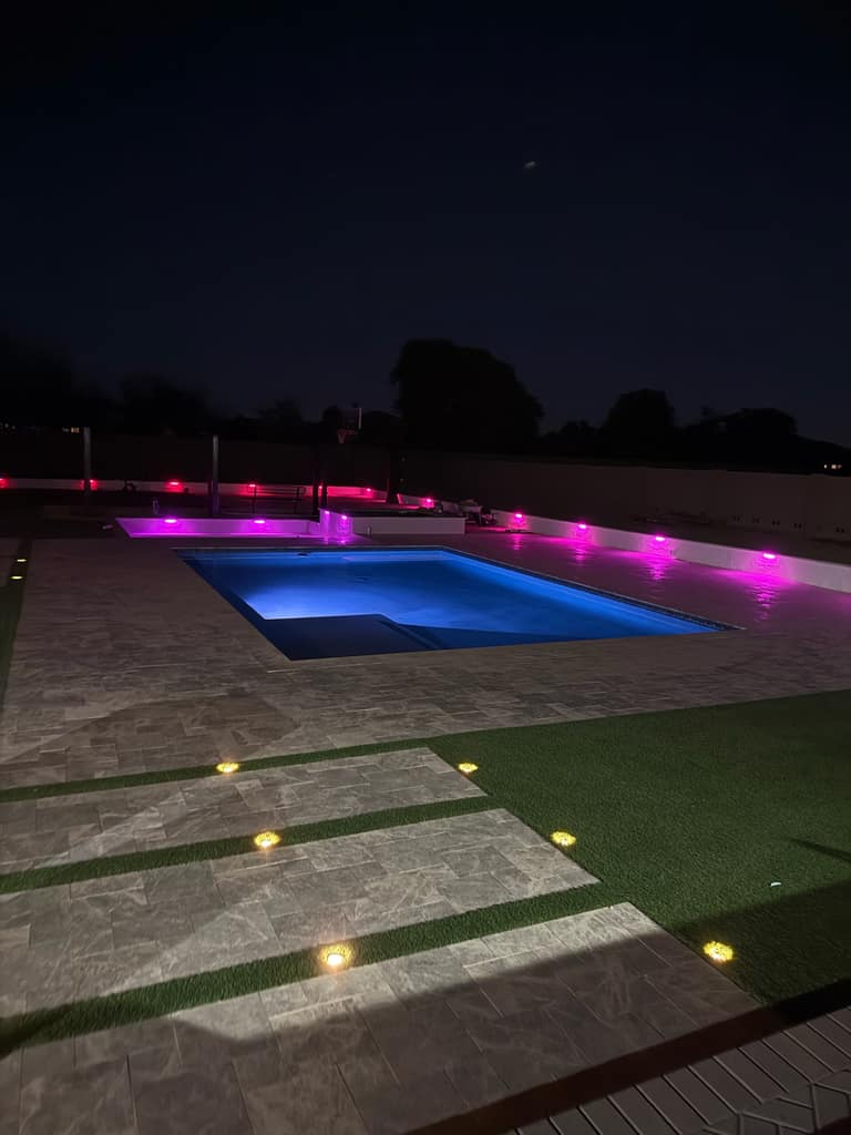 Rectangle pool with blue LED, pink accent wall lights, travertine deck, and turf-and-paver ribbon walkway at night