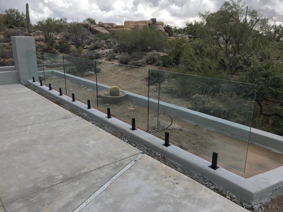 Glass fence panels on black spigots along a poured concrete curb at a desert pool deck.