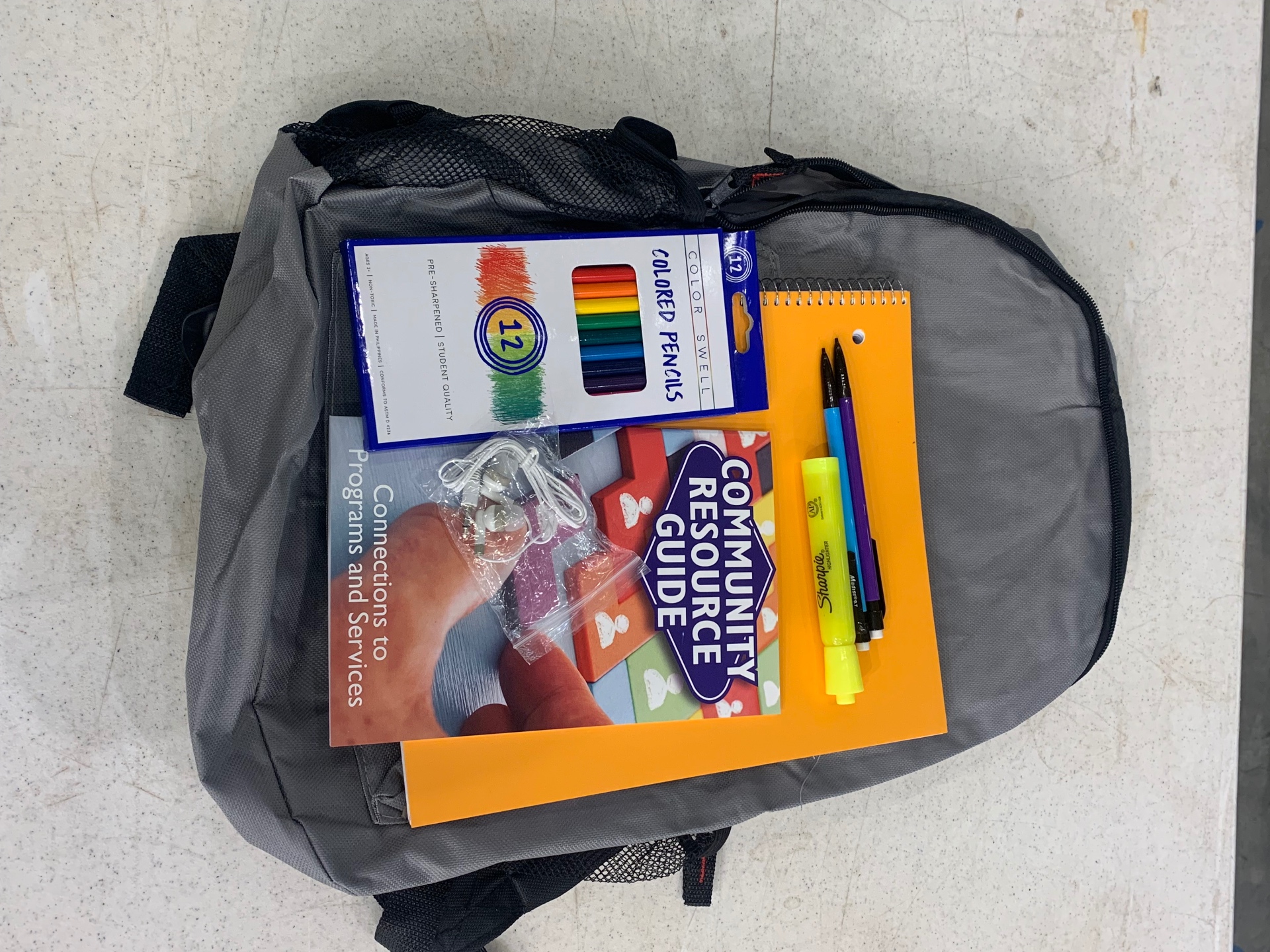 Open backpack showing colored pencils, notebook, pens, highlighter, earbuds, and a Community Resource Guide inside