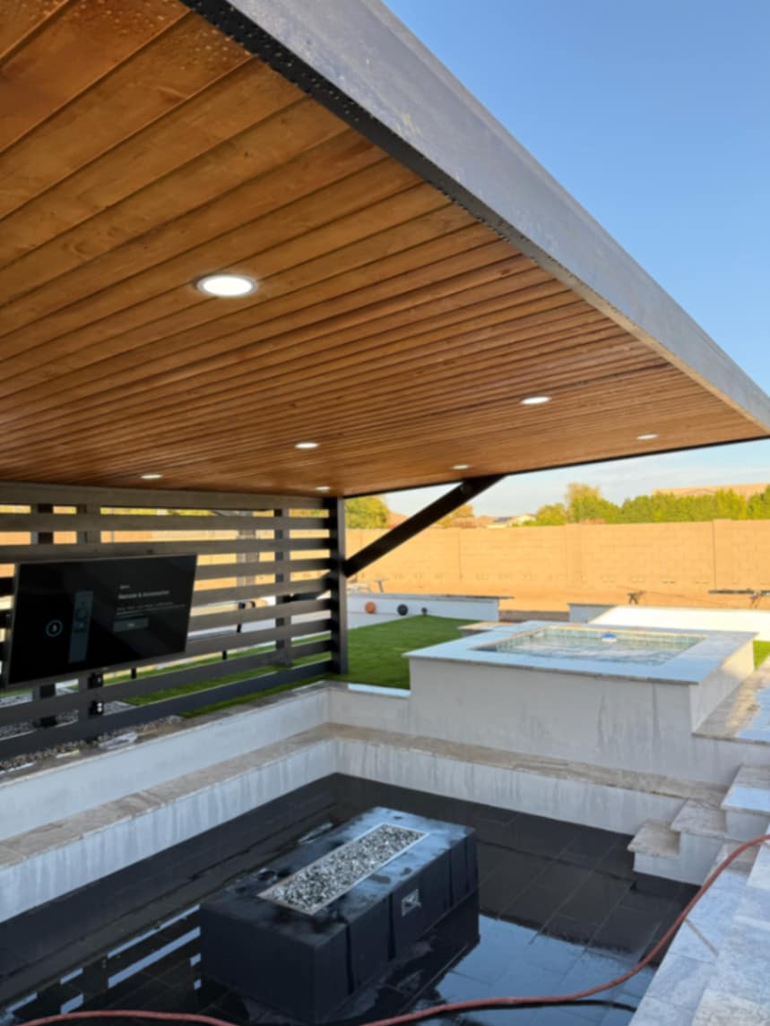 Black pergola sheltering mounted TV and sunken fire lounge with raised spa beyond