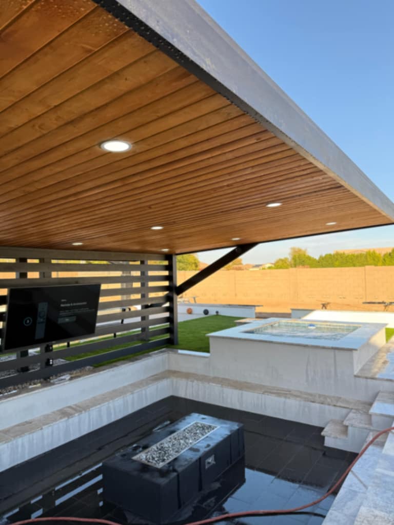 Black pergola sheltering mounted TV and sunken fire lounge with raised spa beyond