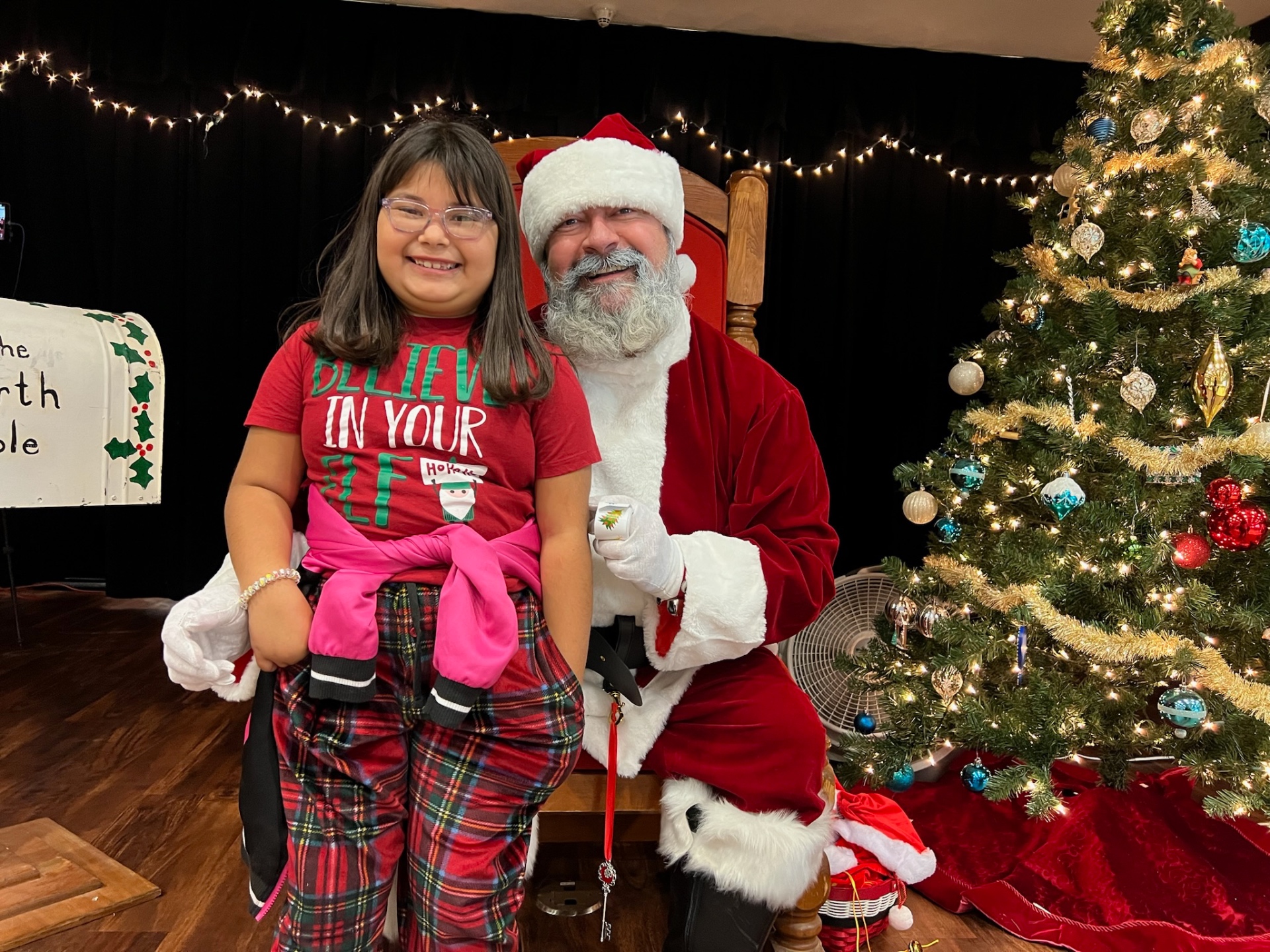 Young girl in a Believe In Your Elf shirt smiling with Santa at Shop With a Cop
