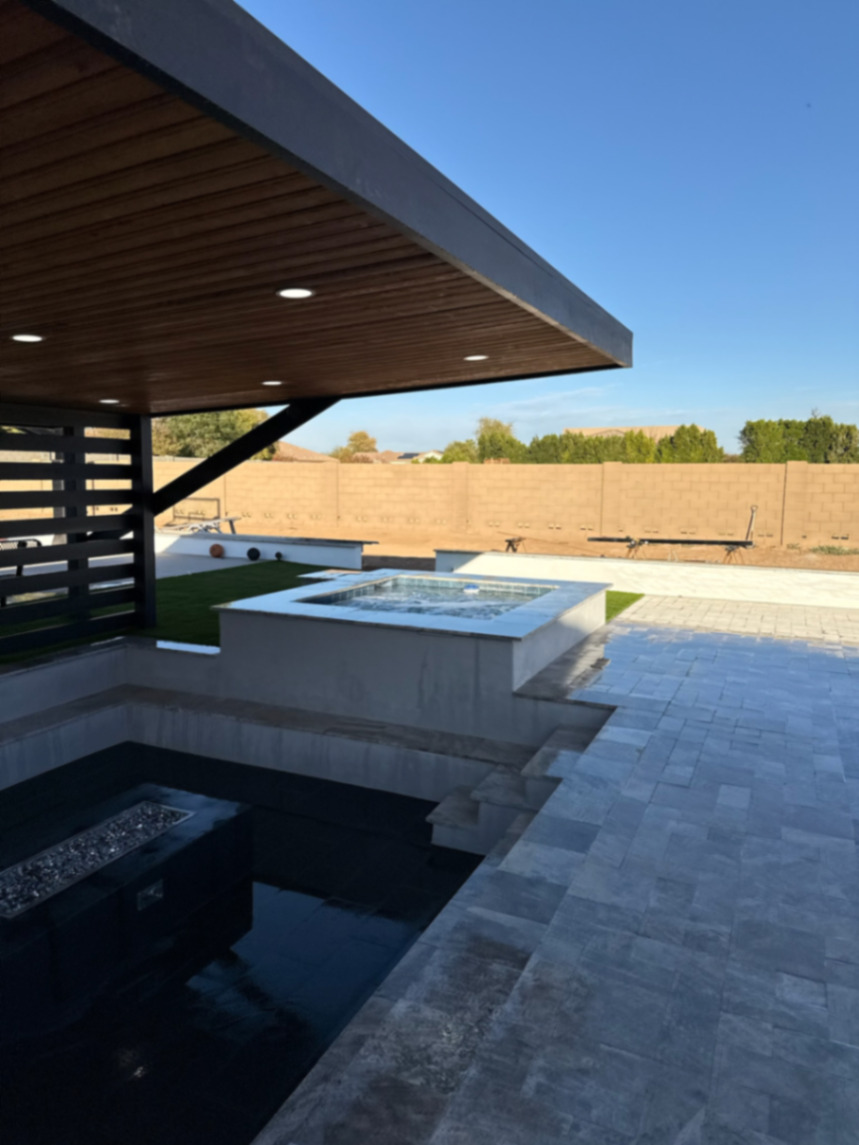 Sunken fire lounge with linear fire pit, black-tile floor, raised spa, and travertine paver deck under a black pergola.