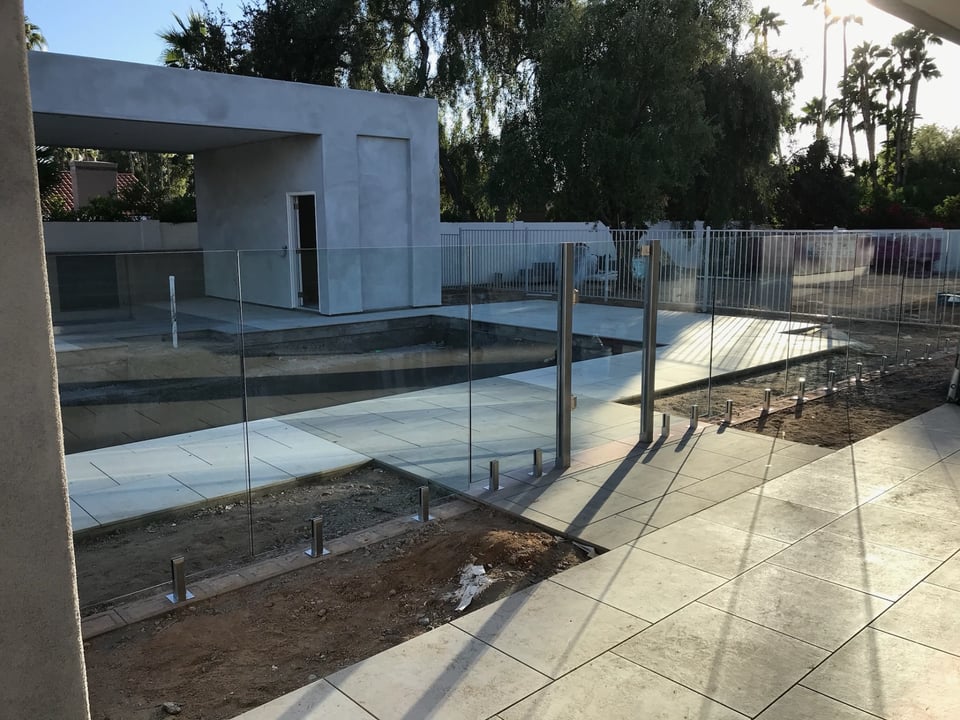 Wide view of glass fence panels around a pool, spa, and pavilion, beside paver decking, with panels on spigots.