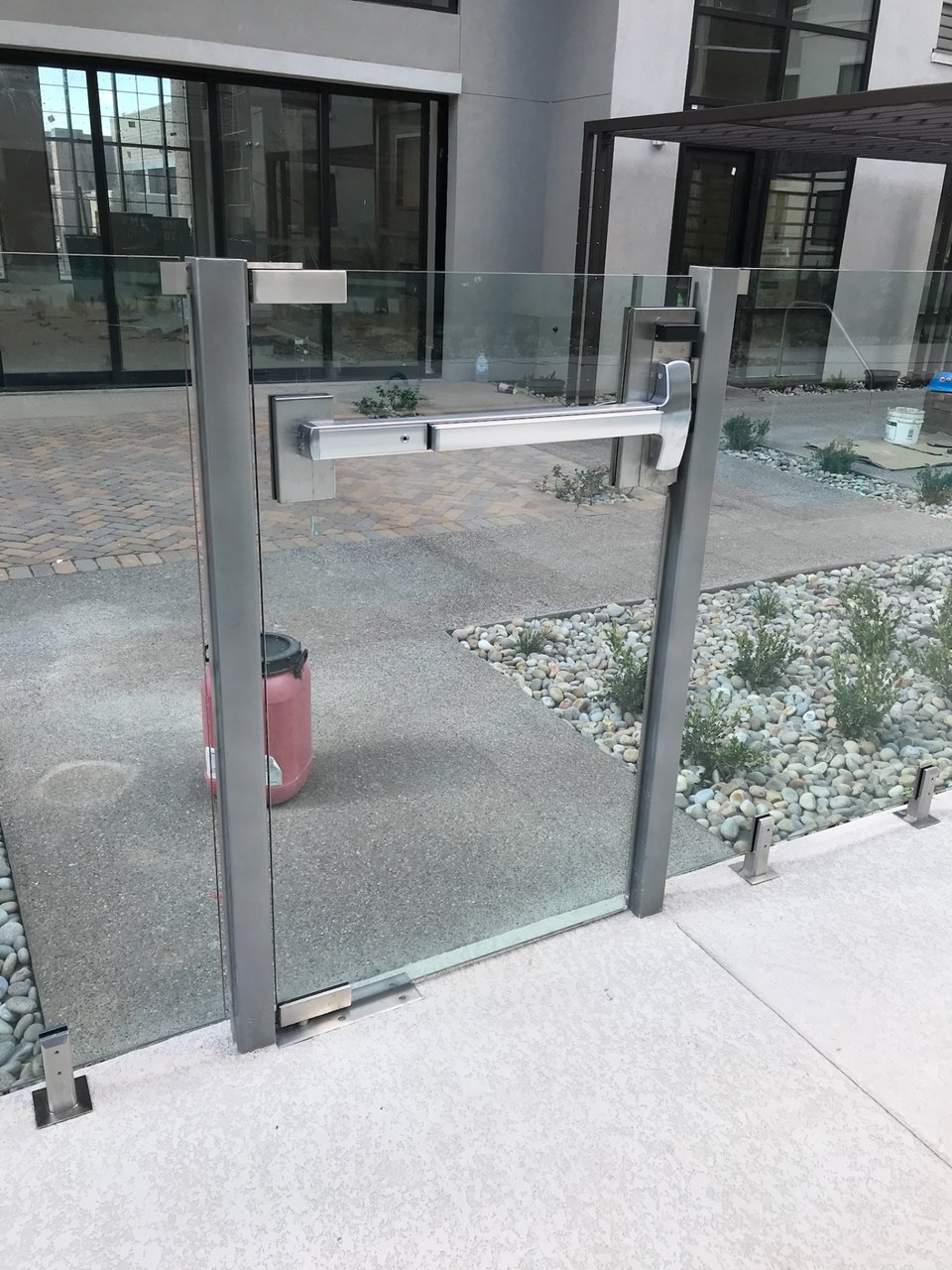 Close view of a metal-framed glass gate with a horizontal push bar, next to adjacent glass fence panels.