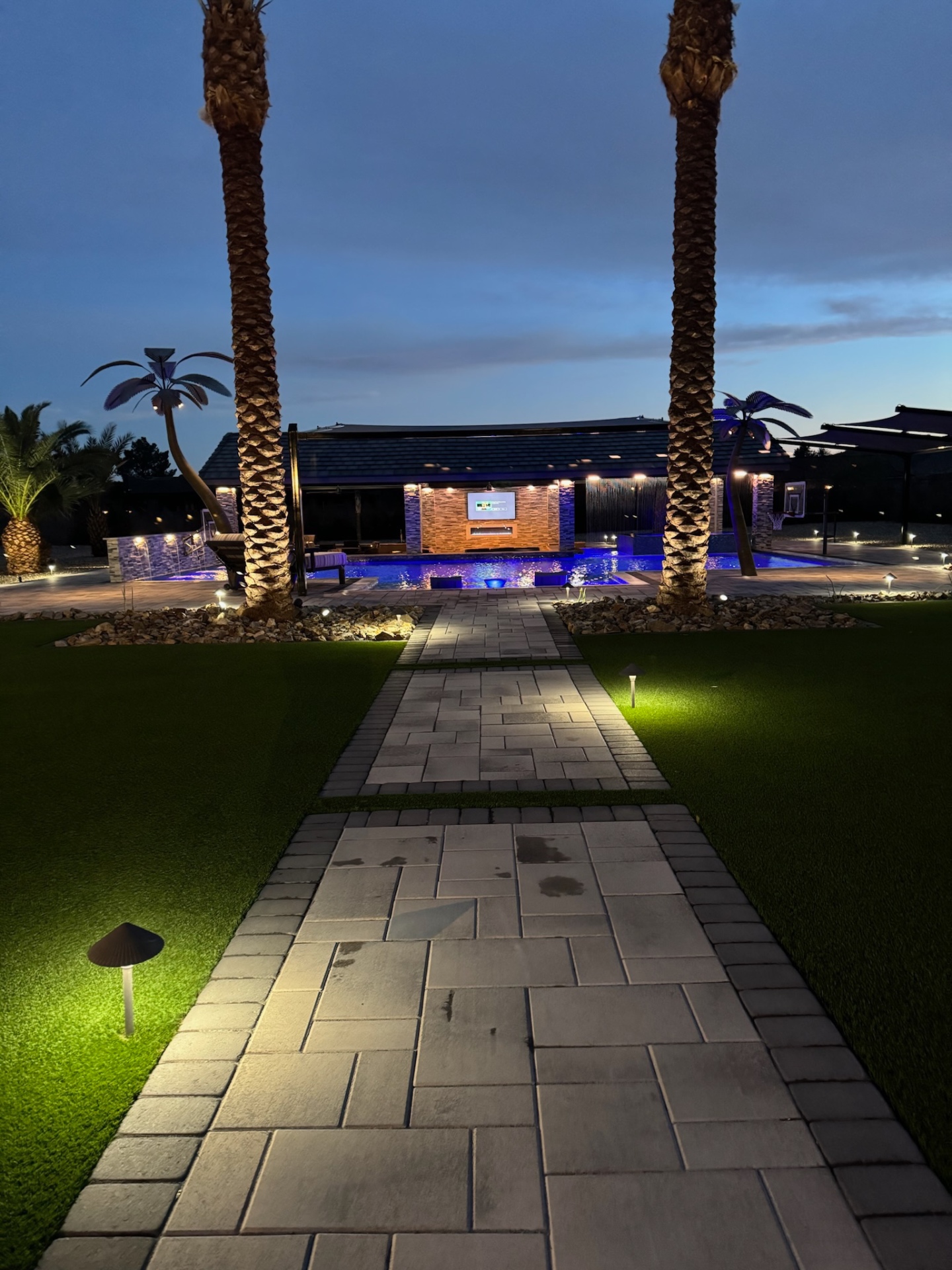 Night entertaining view with lit path, turf, palms, and resort-style pool centerpiece.