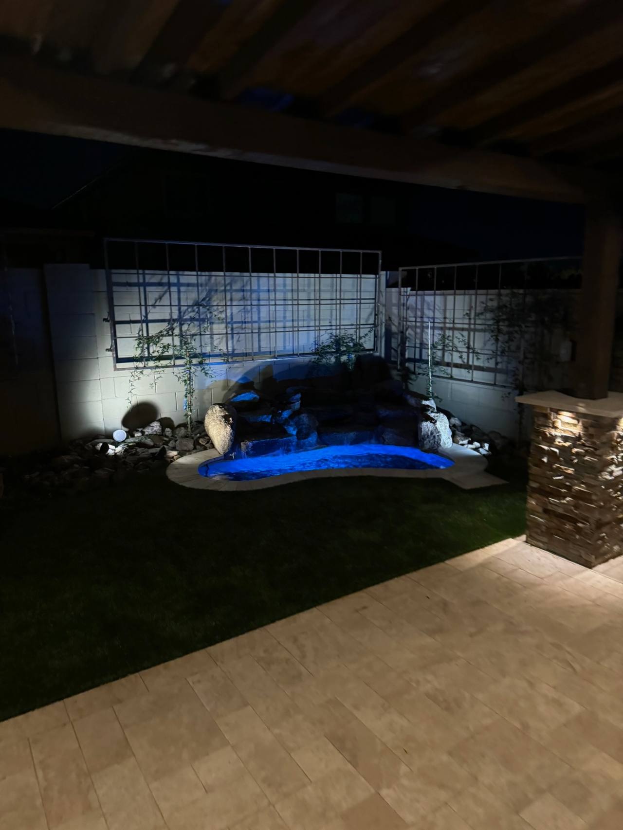 Night view of the freeform spool and boulder waterfall with turf lawn, vine trellises, and stacked-stone patio column