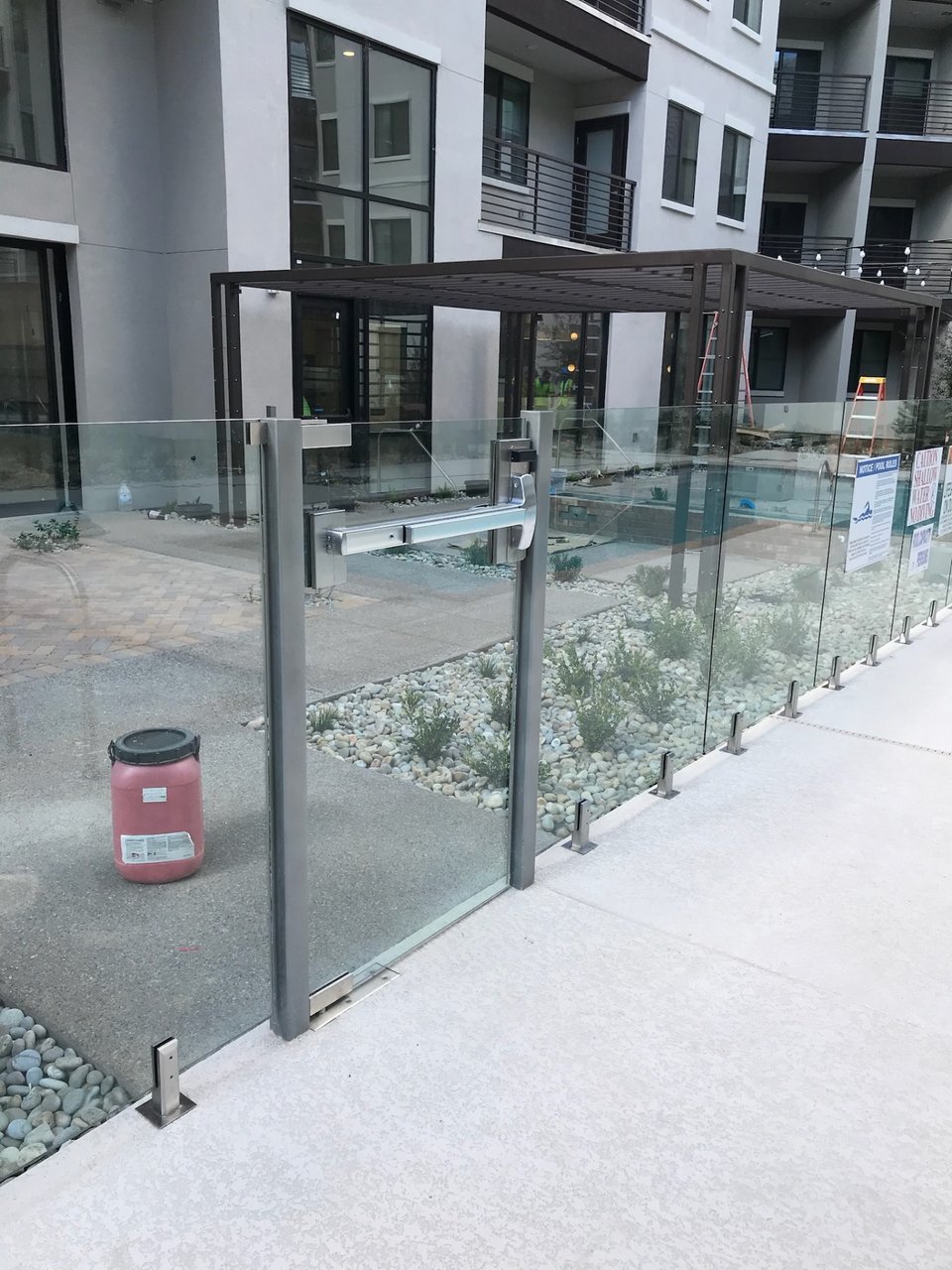 Glass fence gate with a horizontal push bar and a steel shade structure beside a multifamily amenity pool deck.