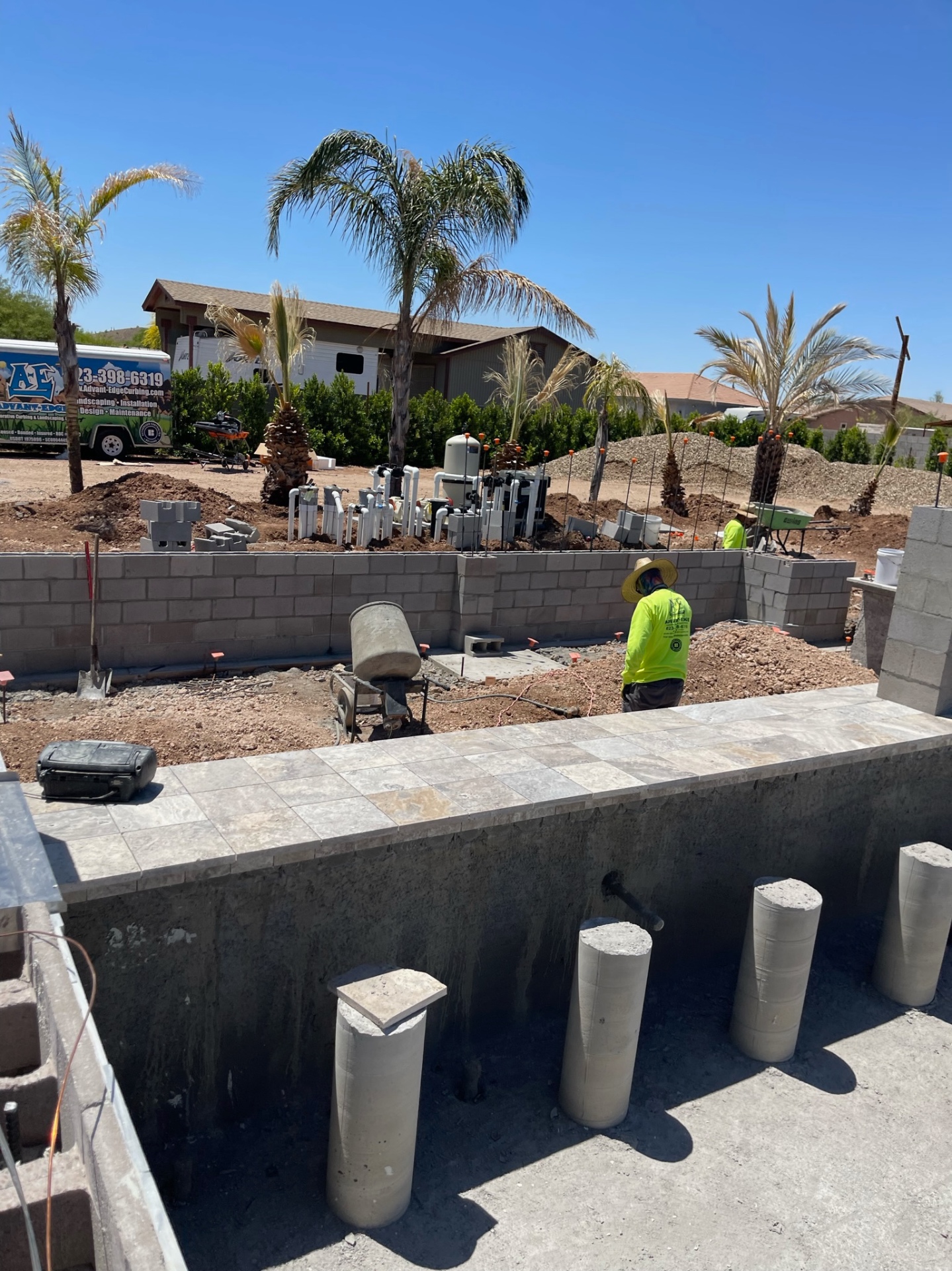 Crew member working beside a raised wall with pool equipment manifold, rough dirt grade, and AE trailer visible behind the shell
