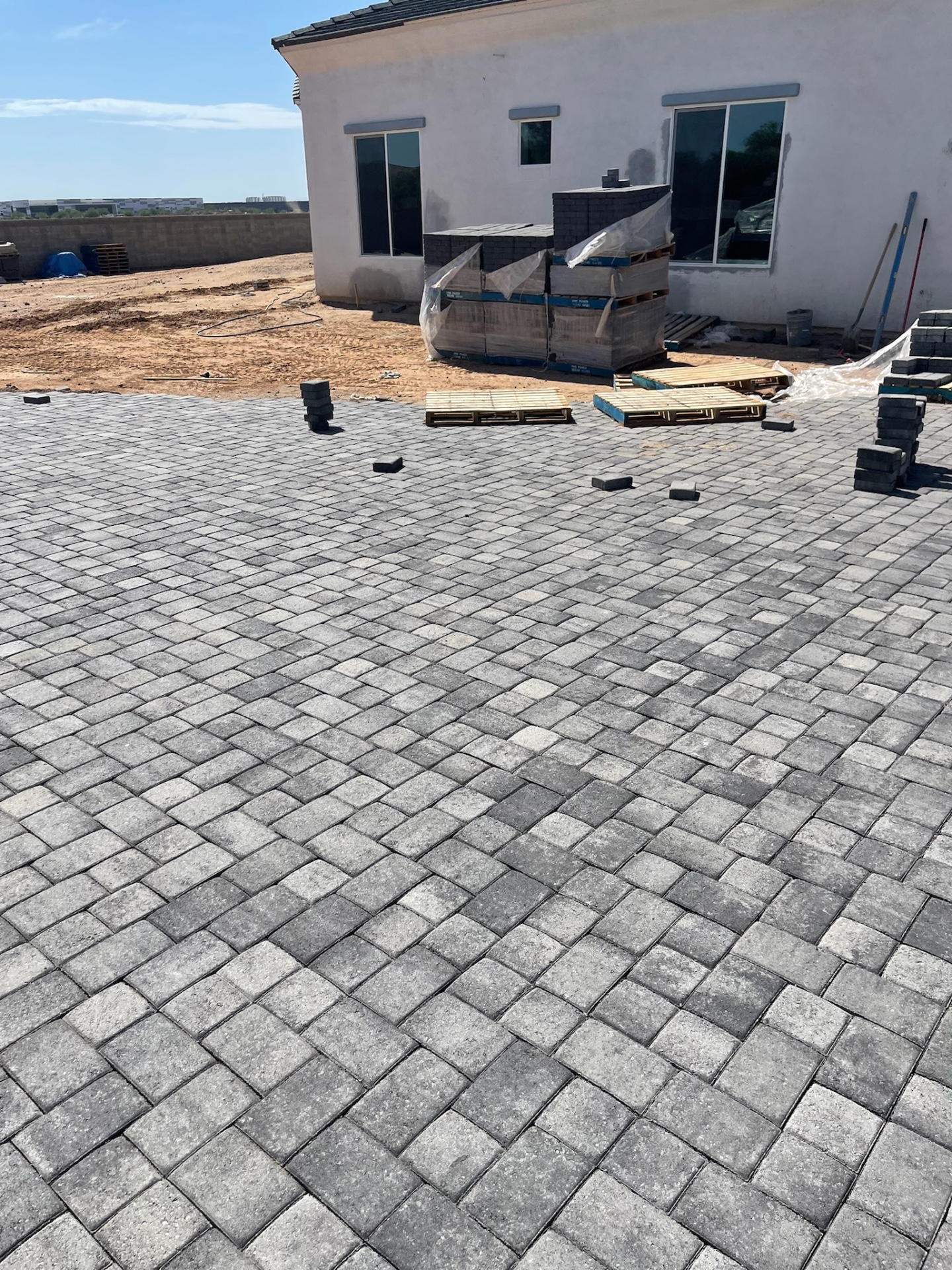 Waddell (Surprise-area) charcoal-blend paver patio nearing completion against rear of stucco home with staged pallets for adjoining runs by AE Outdoor Living