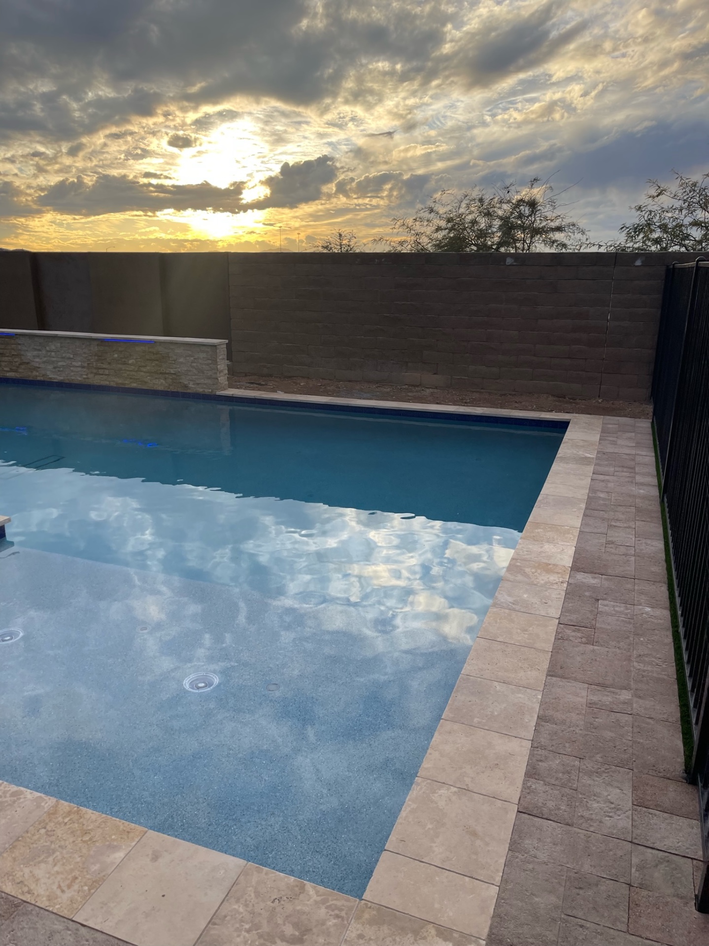 Peoria pool with pebble finish, blue-glass waterline tile, travertine paver deck and black mesh pool safety fence at sunset