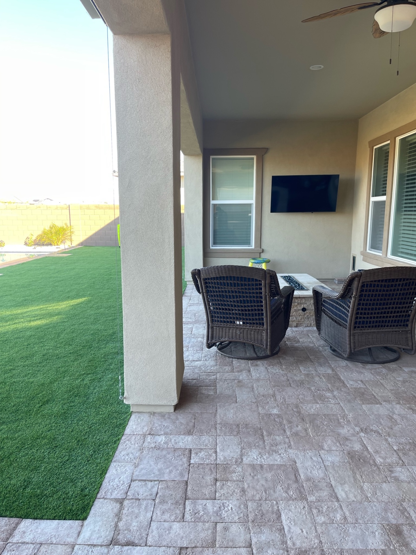 Peoria covered paver patio with wicker swivel chairs, wall-mounted TV and linear fire feature beside artificial turf lawn by AE Outdoor Living