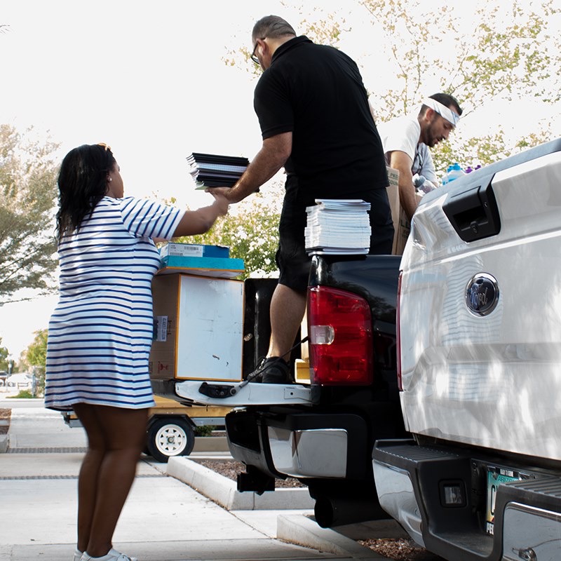 Volunteers loading stacks of notebooks and boxes from a hand truck into a pickup bed