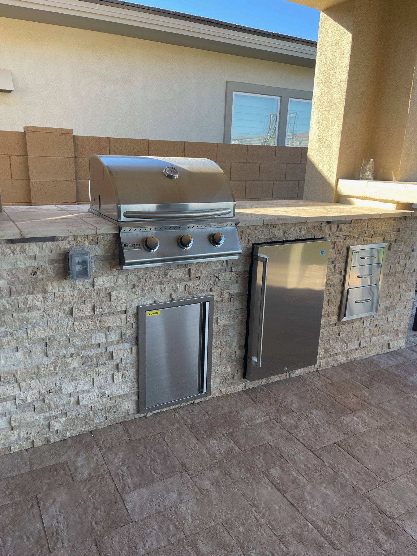 Peoria stacked-stone outdoor kitchen with travertine tile counter, built-in Sizzler stainless grill, beverage fridge and stainless drawer stack on paver patio