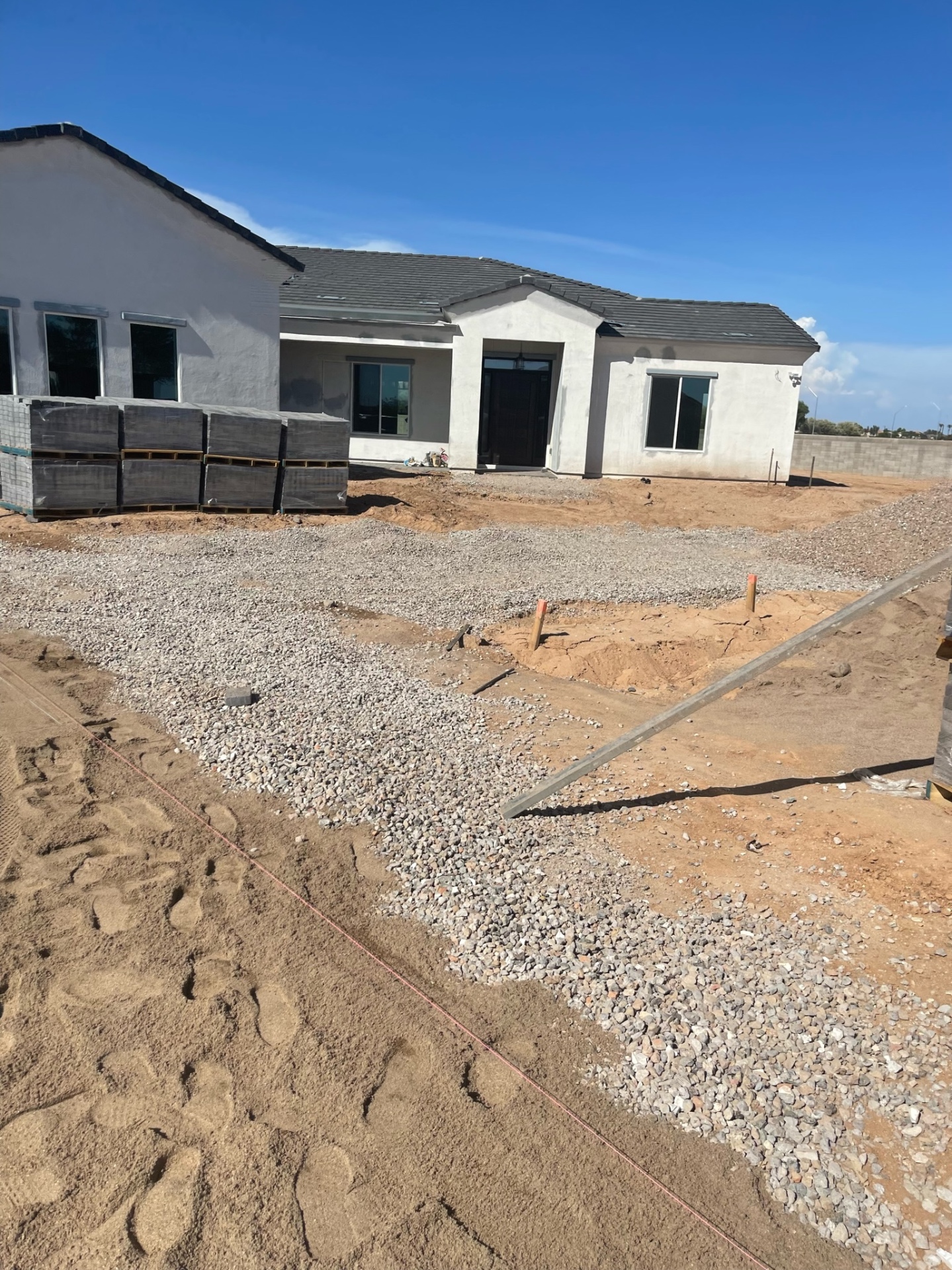 Waddell (Surprise-area) front-yard driveway and walkway prep — compacted ABC base with staged charcoal paver pallets in front of new custom stucco home by AE Outdoor Living