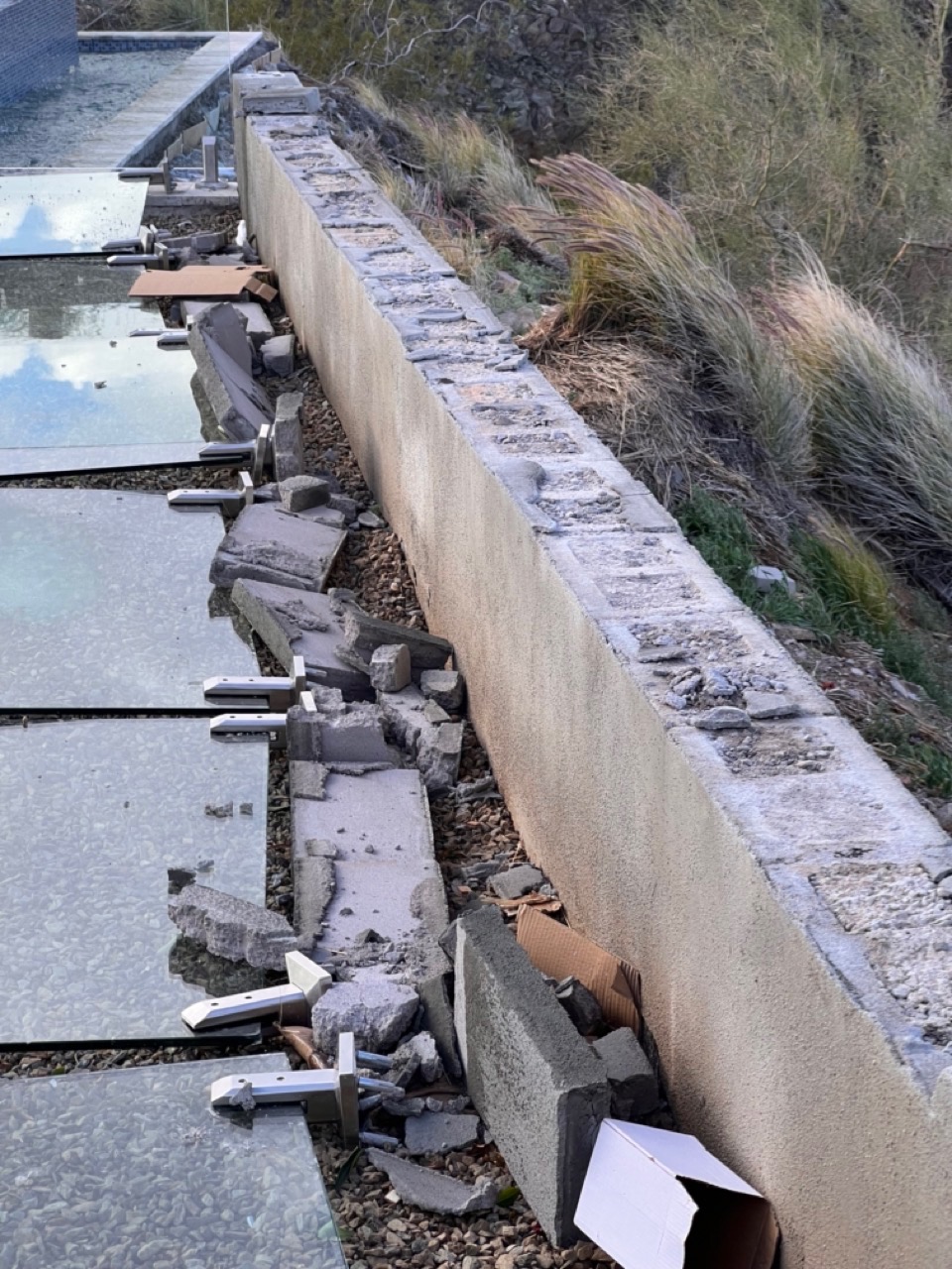Close-up of a crumbled stucco wall cap with bolt-down spigot base plates and torn-out block fragments where a glass pool fence used to be anchored.