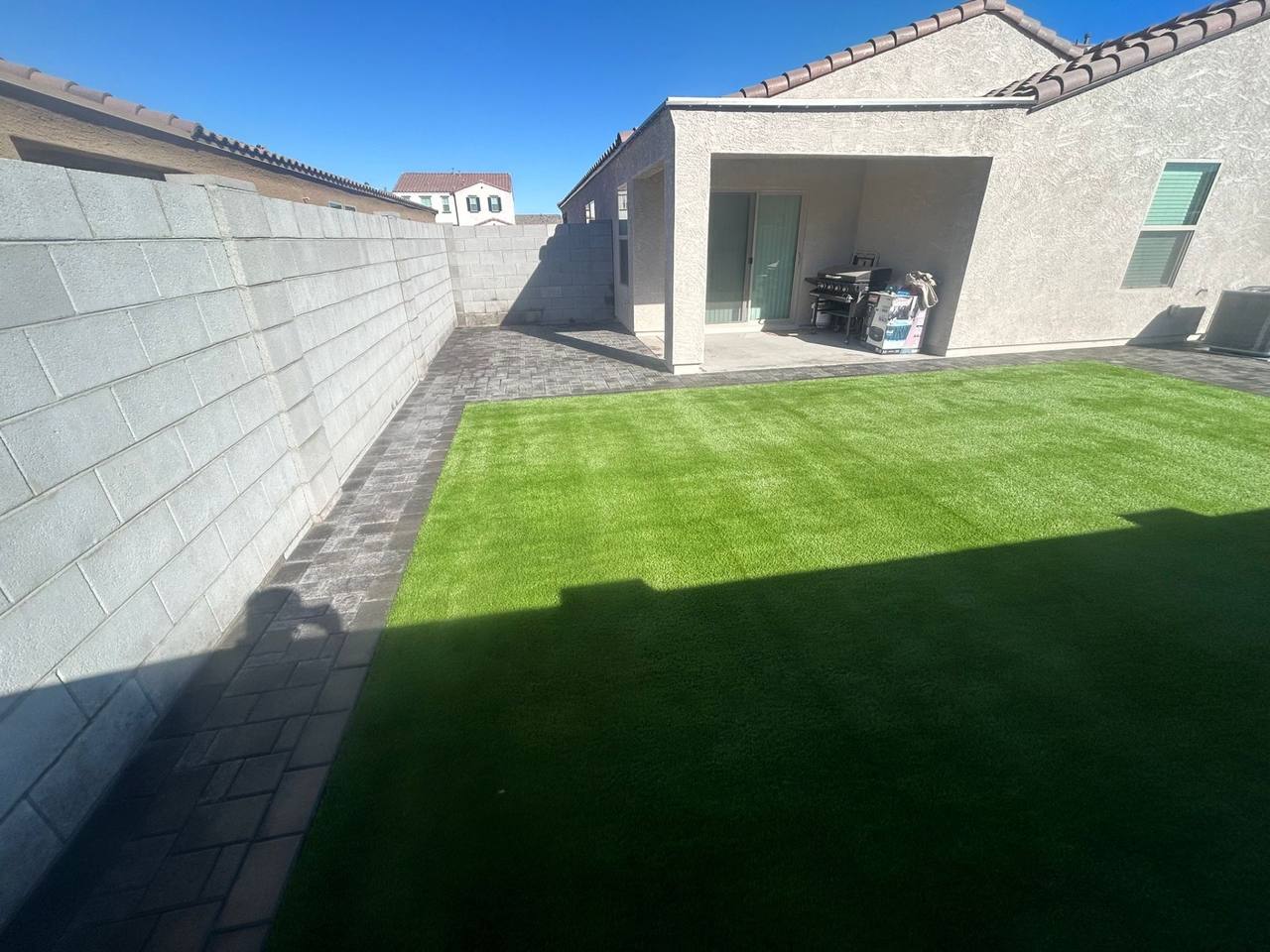 Artificial turf lawn centered inside a charcoal paver patio beside a covered patio