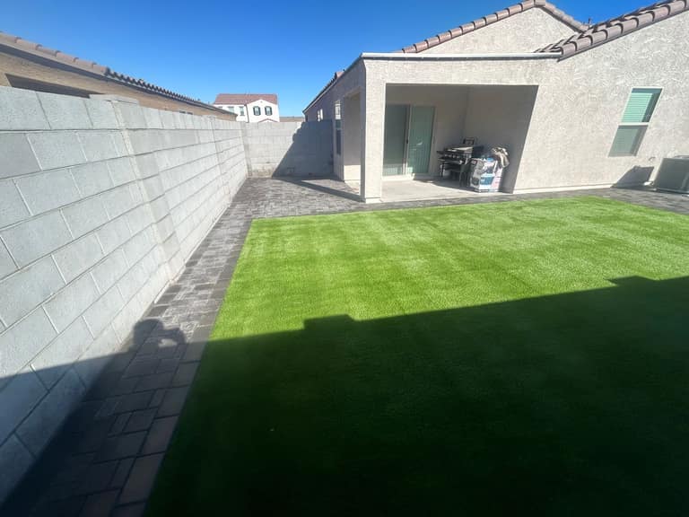 Artificial turf lawn centered inside a charcoal paver patio beside a covered patio