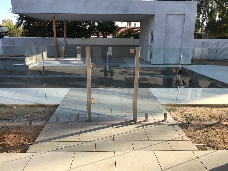 Glass fence panels and a gate between brushed metal gate posts, beside a paver-finished pool deck.