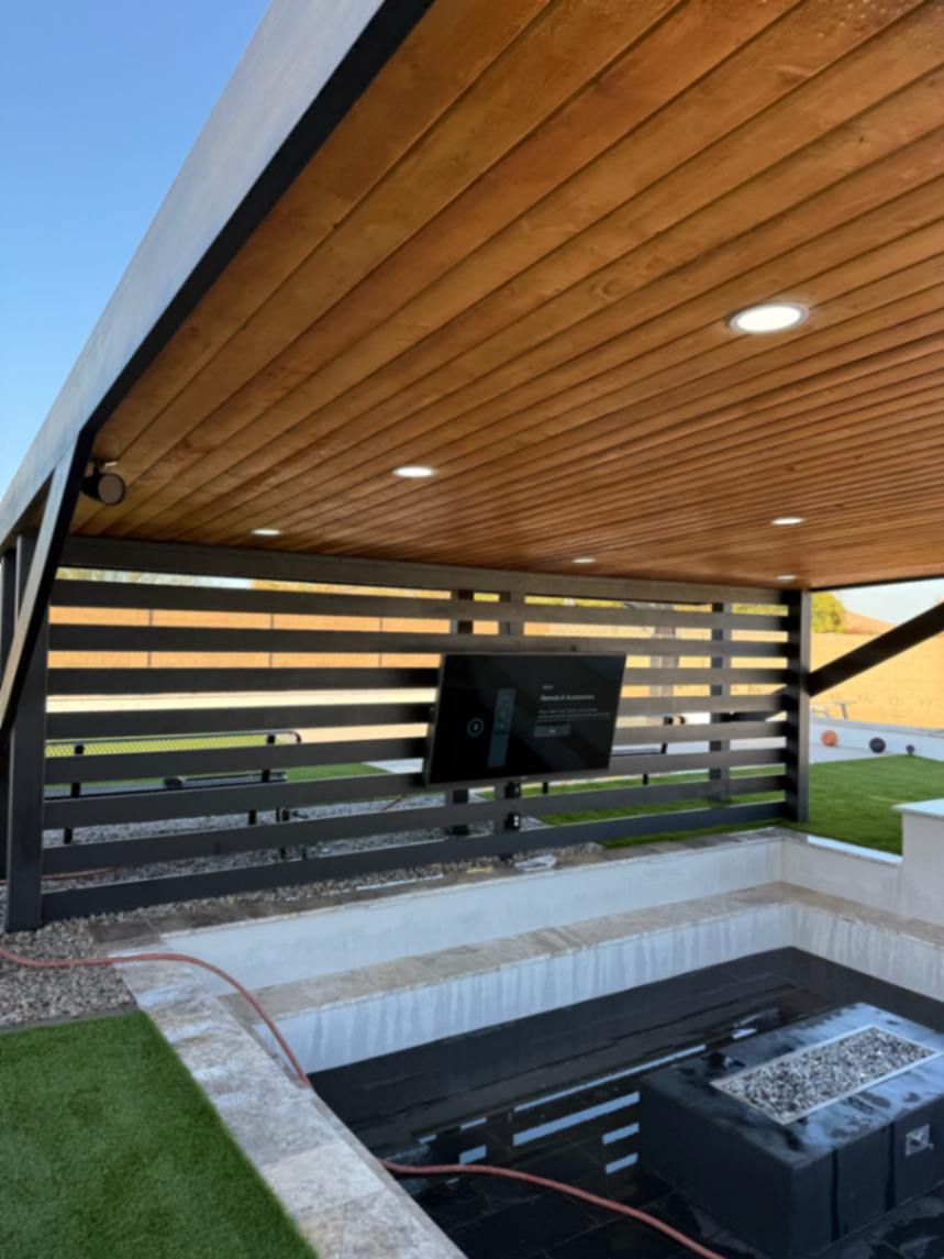 Thorn residence, Mesa AZ — sunken lounge with black linear fire pit under a cedar-plank covered patio, horizontal steel slat privacy wall with mounted TV, and turf edge.