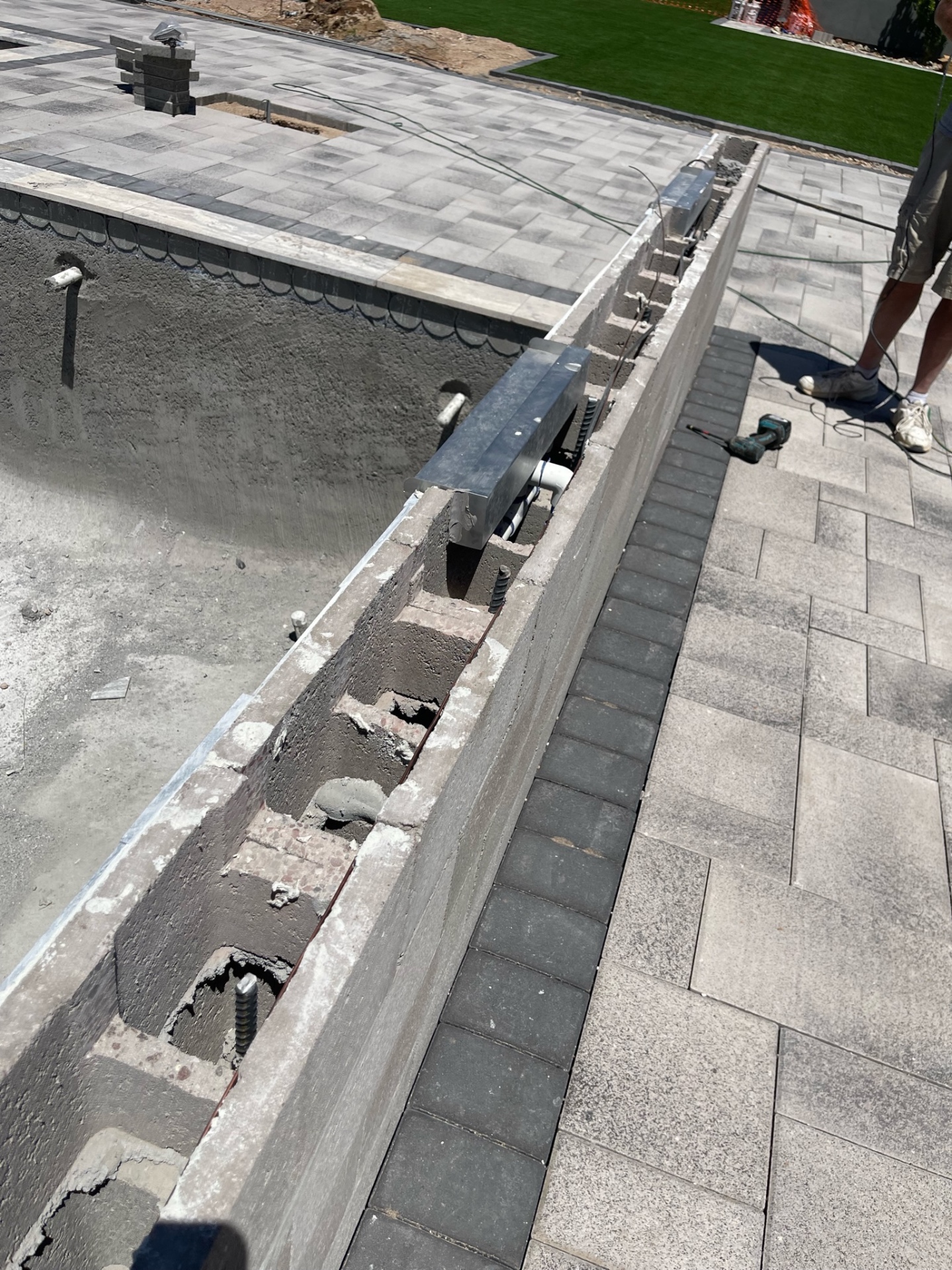 Close-up of a raised wall with metal beam brackets, exposed block cells, dark paver border, and large-format gray deck pavers