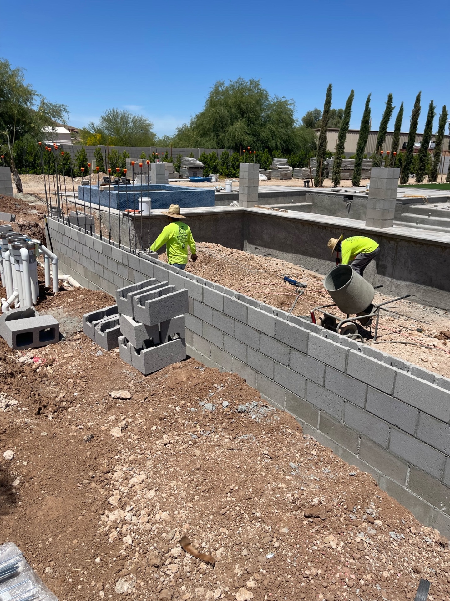 Two AE crew members laying CMU block on a long perimeter wall with vertical rebar, raised blue-tiled spa, and pool shell behind